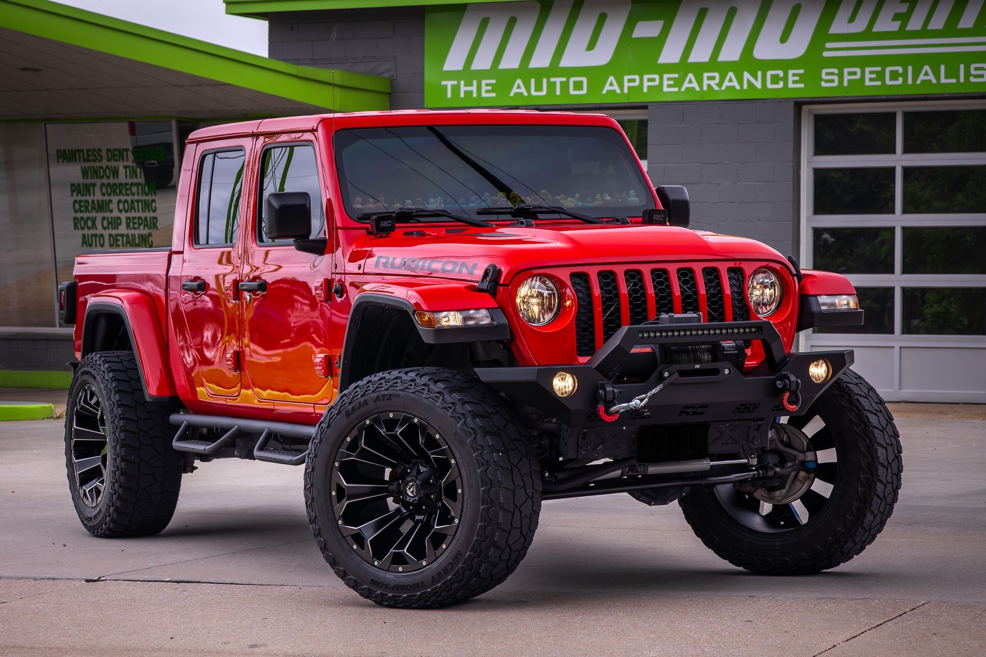 Red Jeep Gladiator with black wheels parked in front of a green sign for an auto appearance shop.
