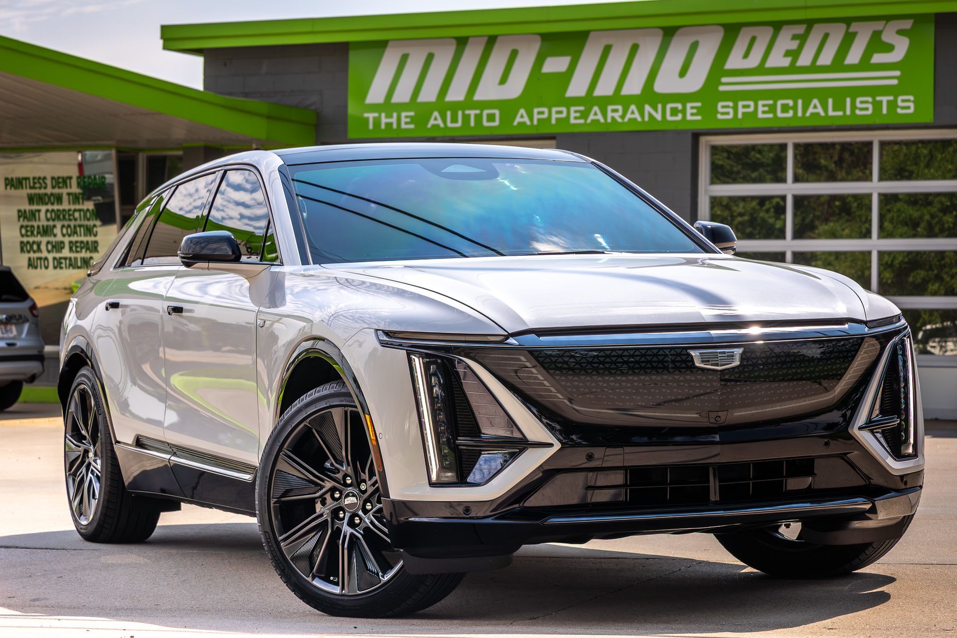 White Cadillac SUV parked in front of a car appearance shop, with black wheels and a green sign.