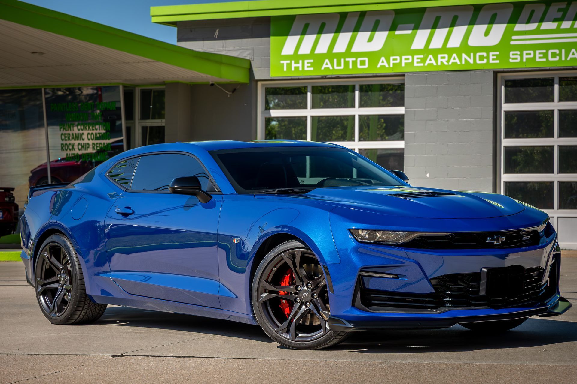 Blue Chevrolet Camaro parked in front of an auto appearance shop with black wheels and red brake calipers.