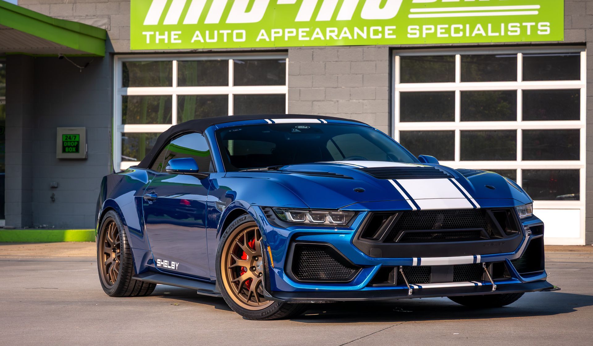 Blue convertible Ford Mustang Shelby GT500 with white racing stripes in front of a shop.