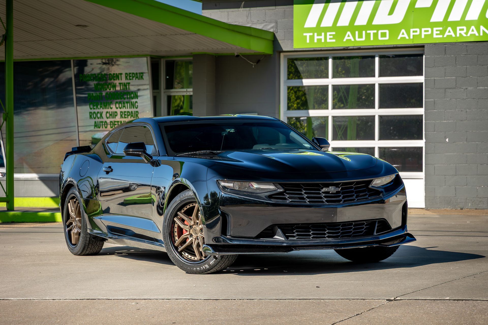 Black Chevrolet Camaro parked in front of an auto appearance shop with bronze-colored wheels.