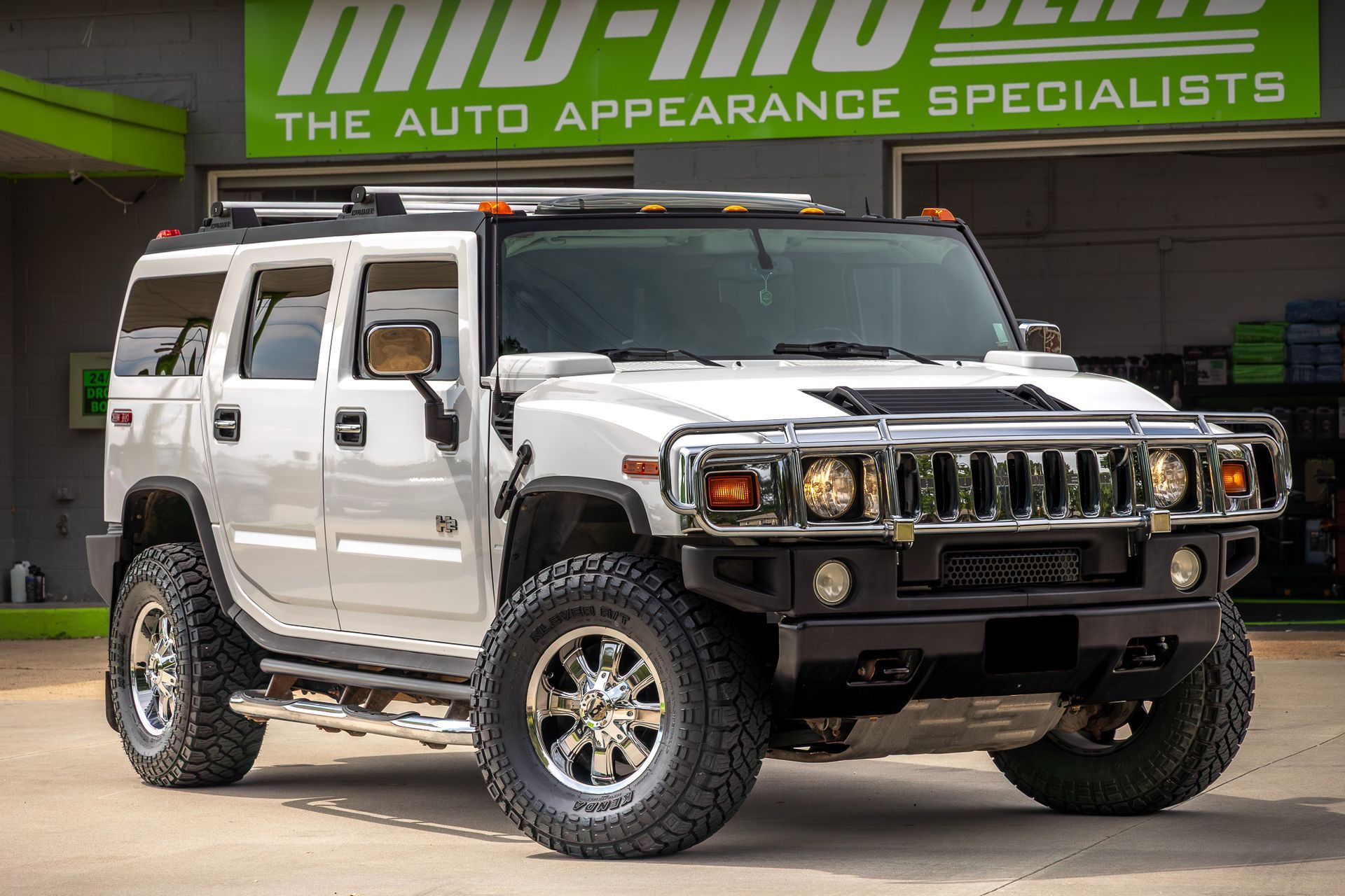White Hummer H2 SUV parked in front of an auto appearance shop with green signage.