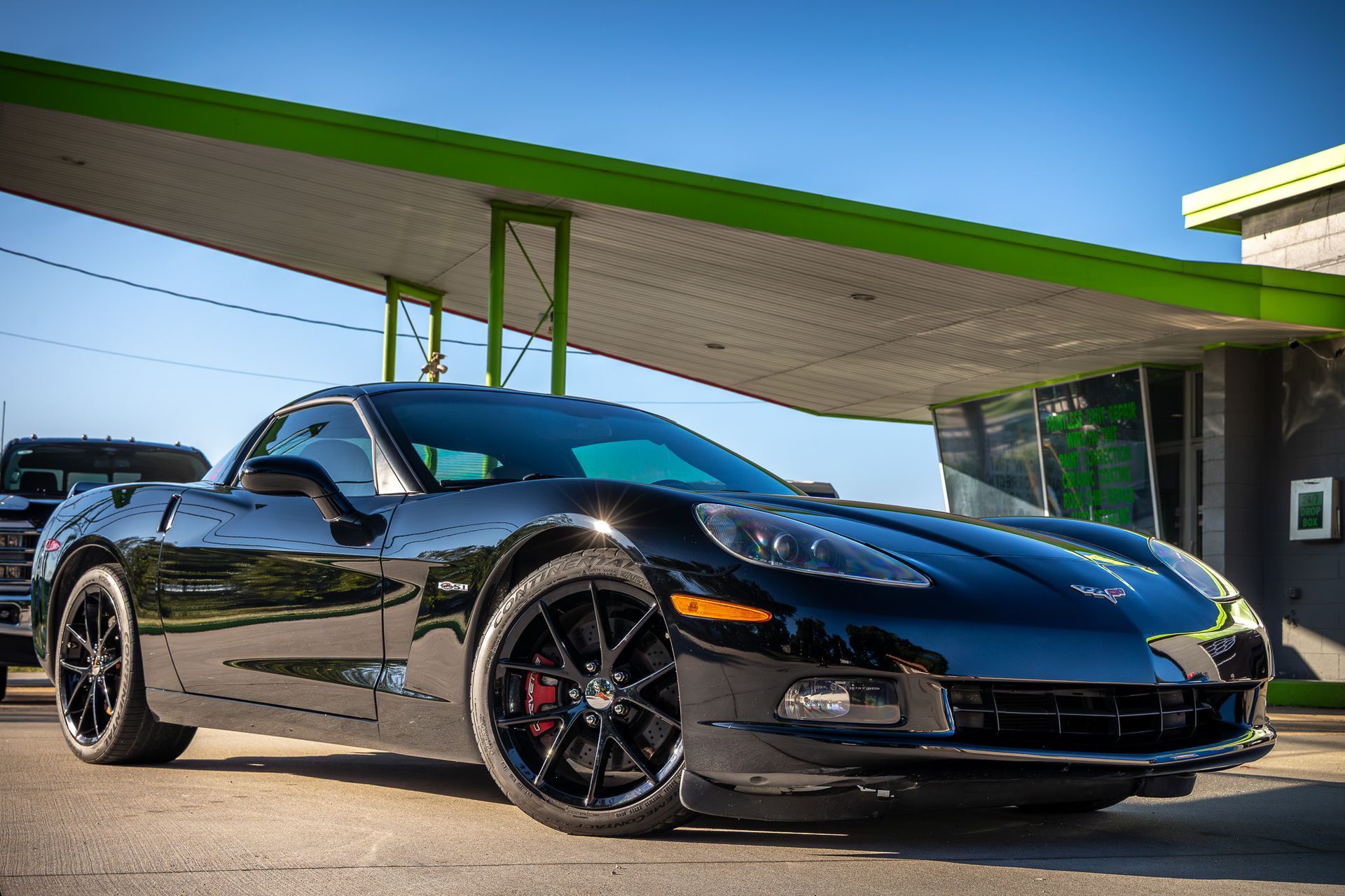 Black Corvette sports car parked under a green-roofed structure, on a sunny day.