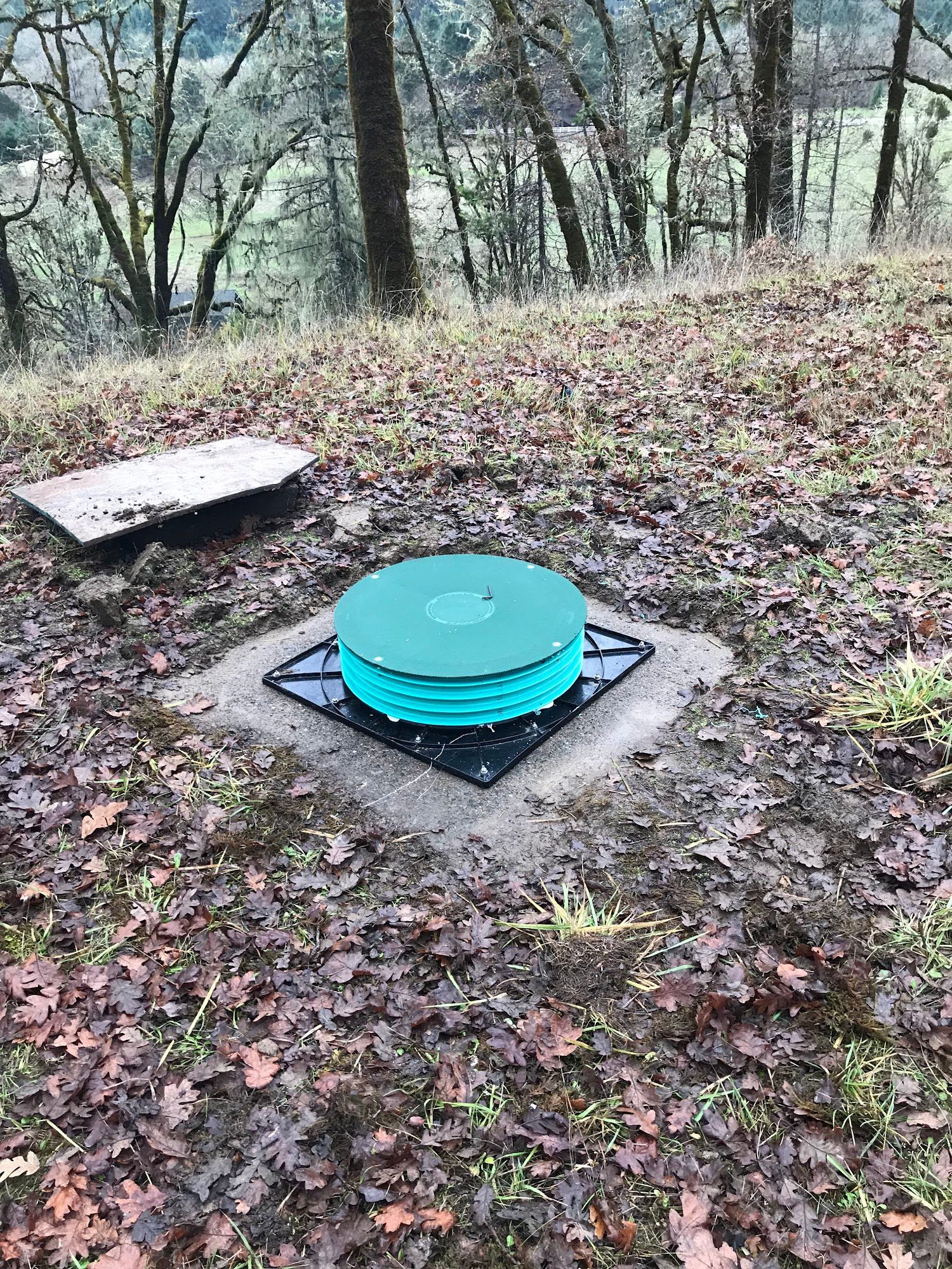 Green septic tank lid set in a square frame on a grassy hillside, next to a concrete cover.