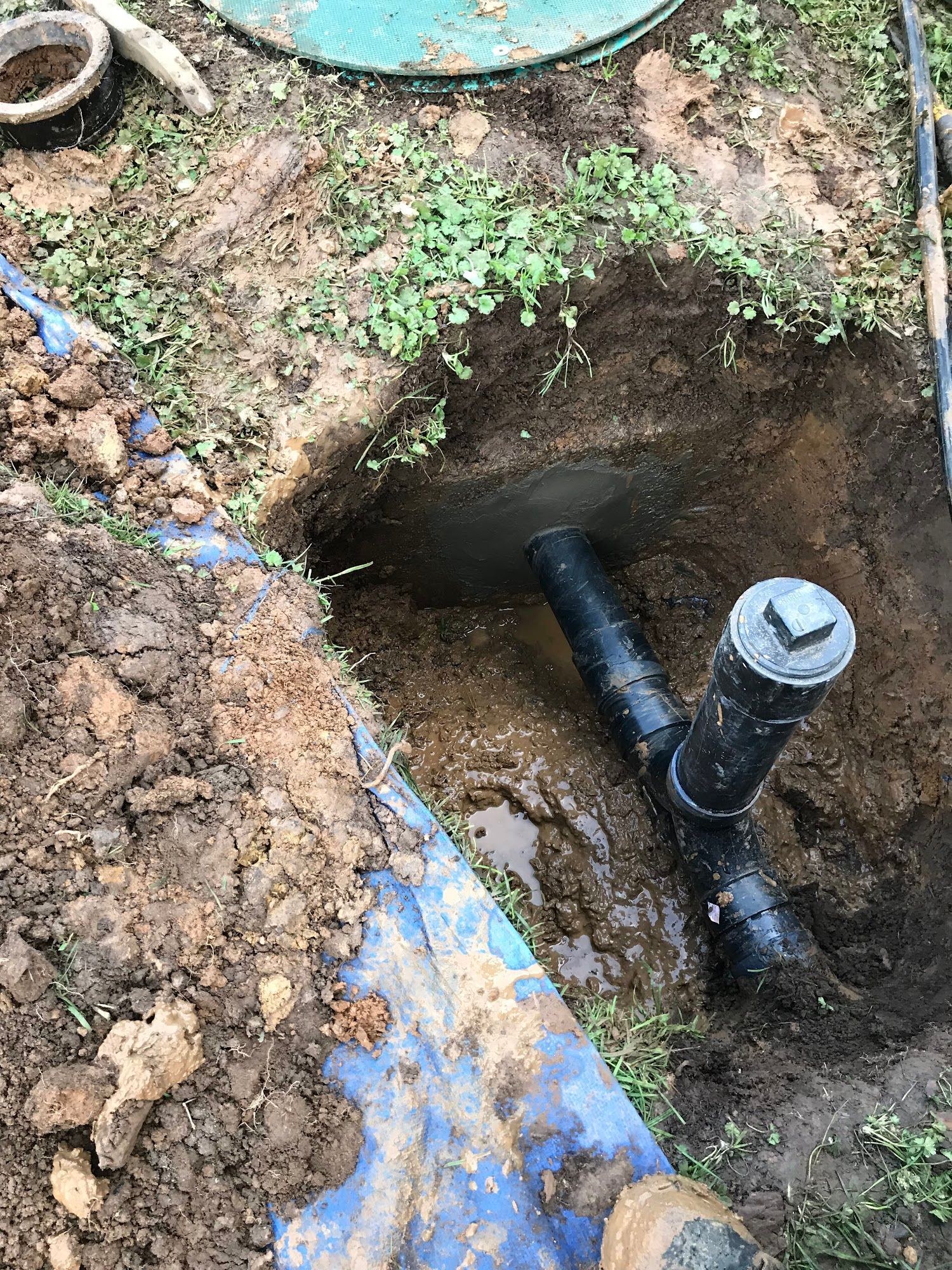 Black pipes and a T-fitting in a muddy excavated pit, likely for plumbing or sewage.