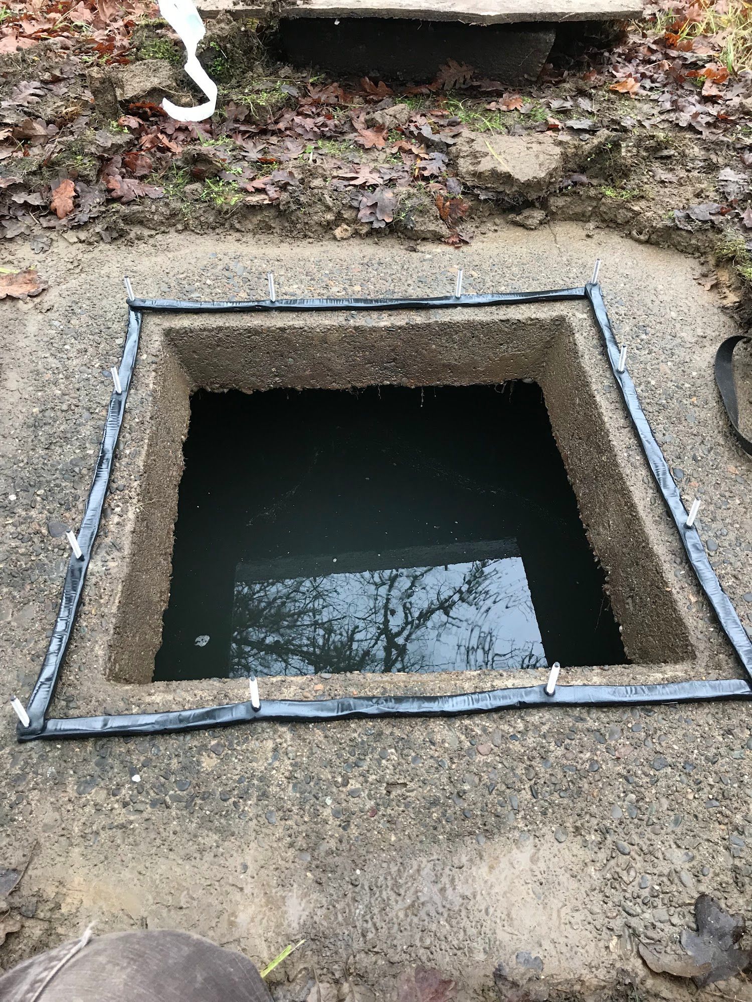 Open square pit filled with water, surrounded by concrete and dark edging, in an outdoor setting.
