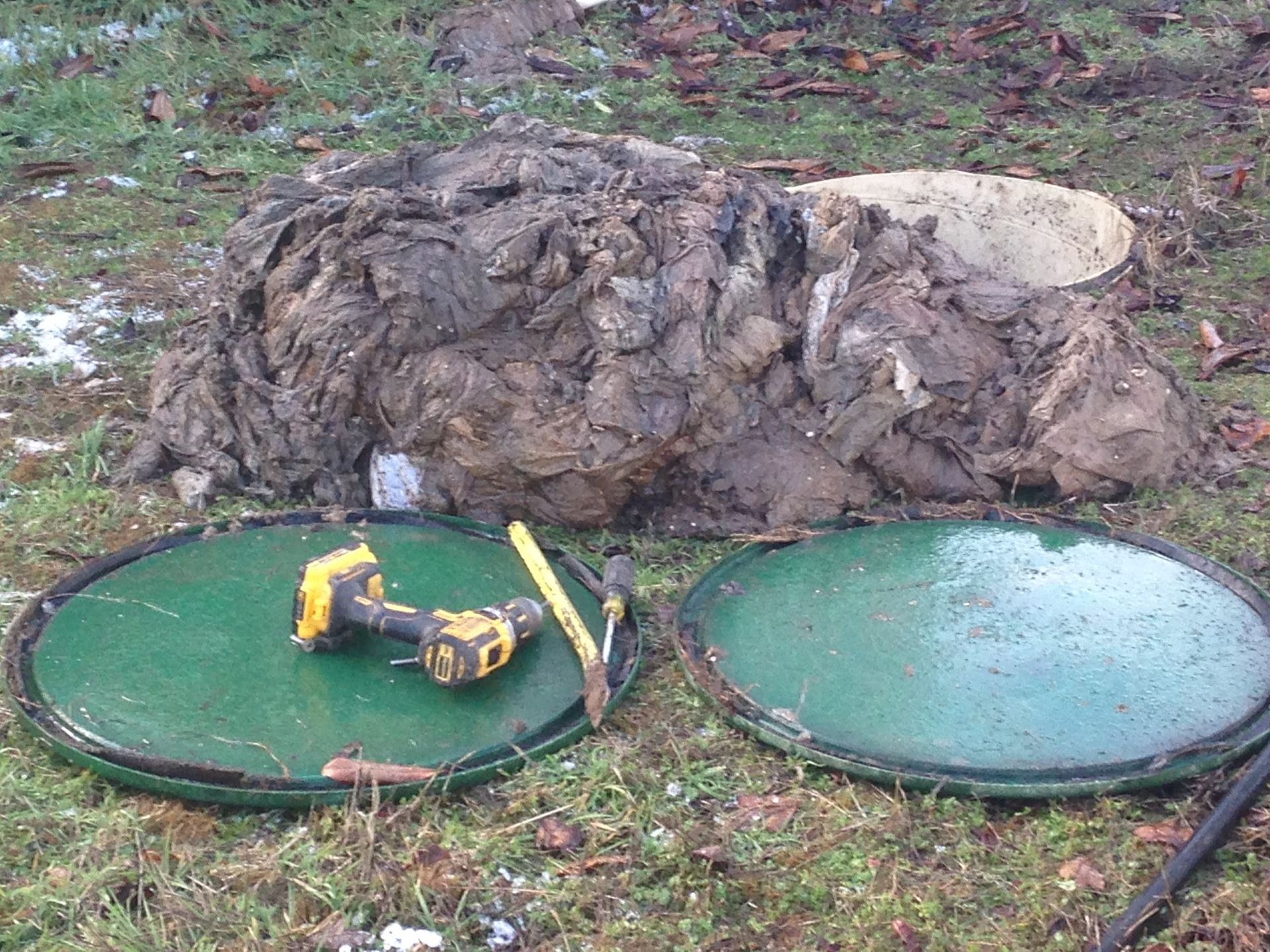Opened septic tank covers with debris pile on grass; power drill and tool rest on one cover.