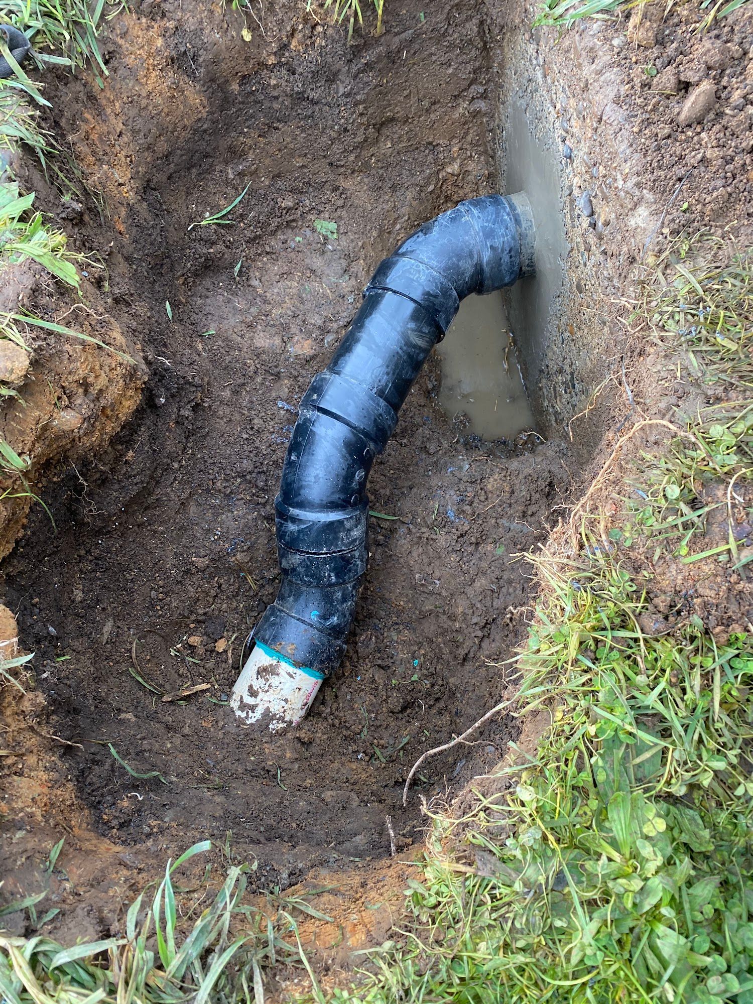 Black corrugated drain pipe in a dirt trench, connected to a concrete wall.