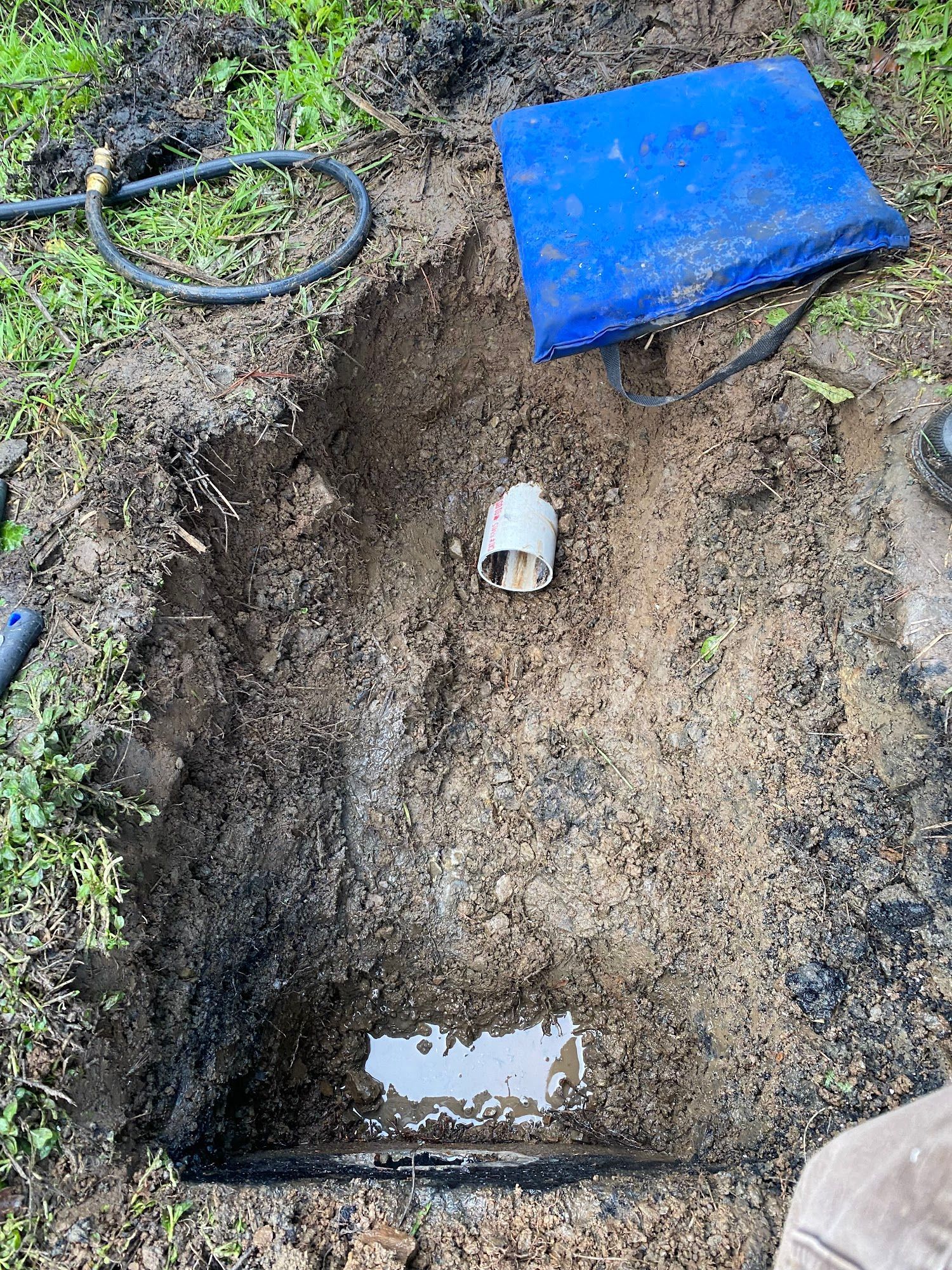Hole dug in muddy ground with white pipe and blue tarp; hose lies nearby.