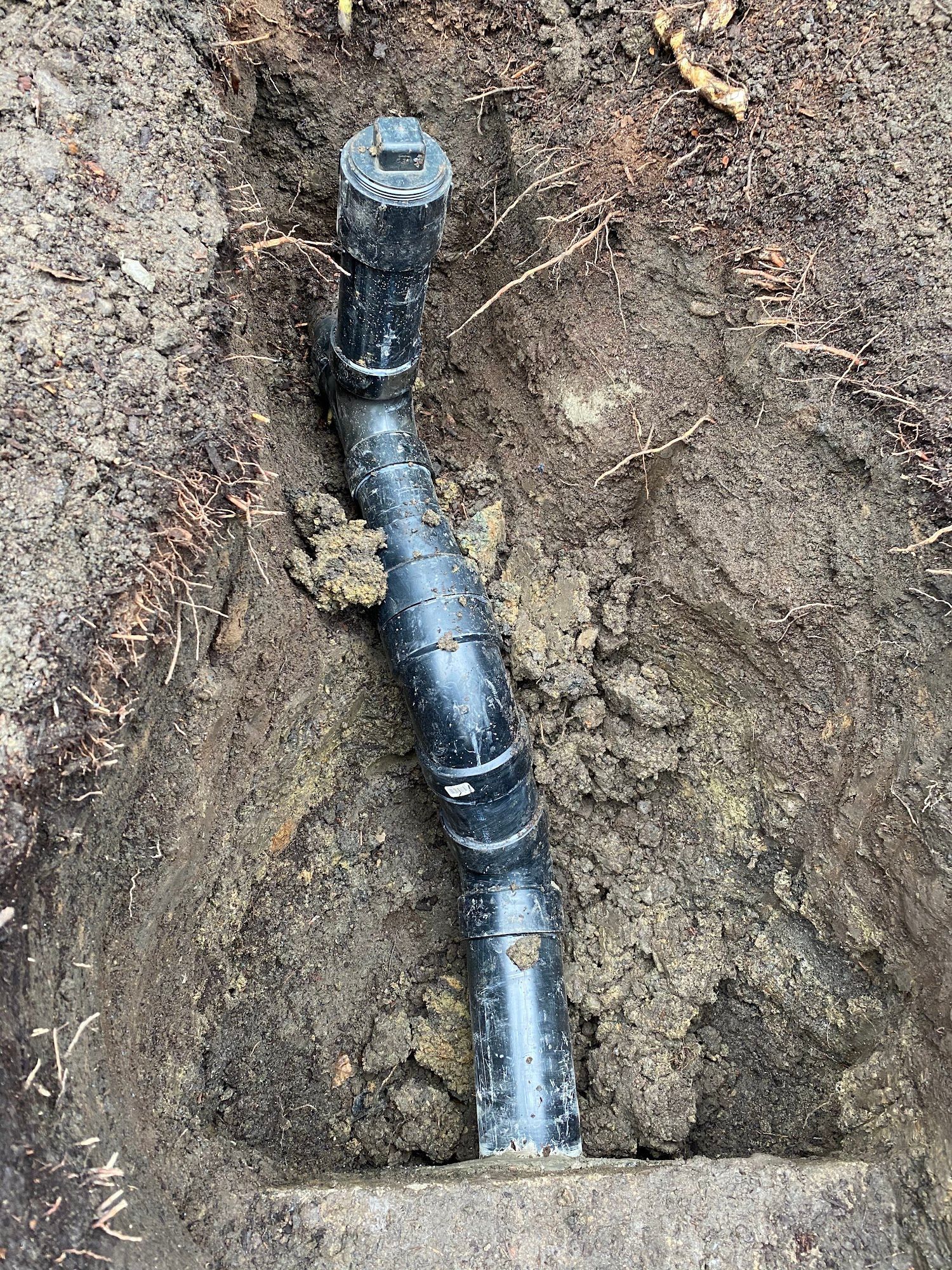 Black pipe section in a dirt trench, possibly part of a plumbing or drainage system.