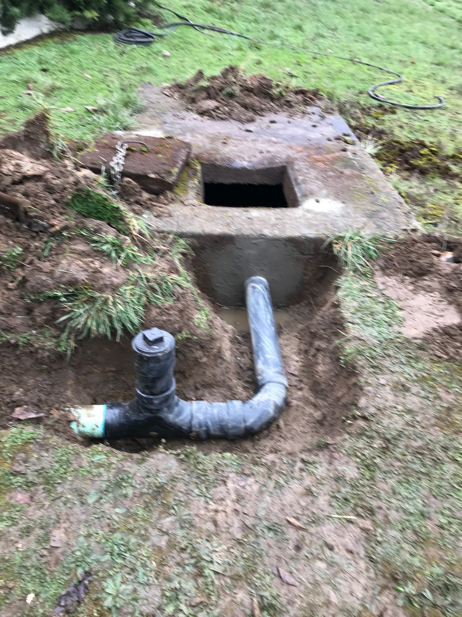 Open concrete box with exposed black pipes in a grassy area, possibly part of a septic system.
