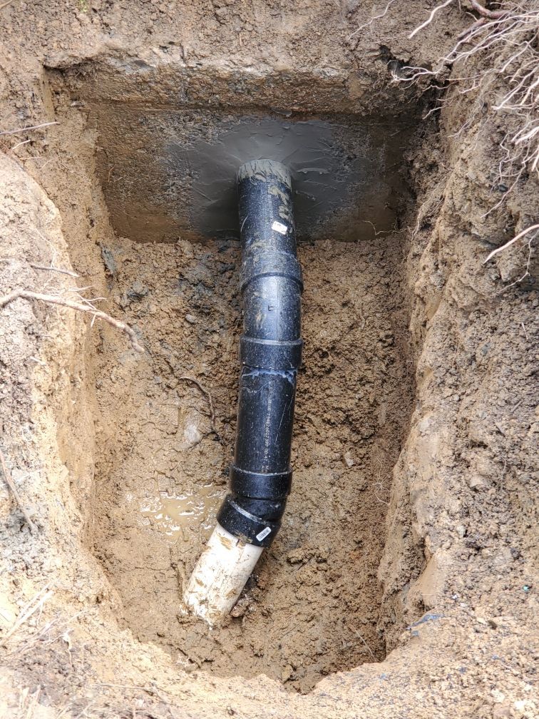 Black pipe emerging from concrete, buried in a rectangular soil trench. The bottom pipe is white.