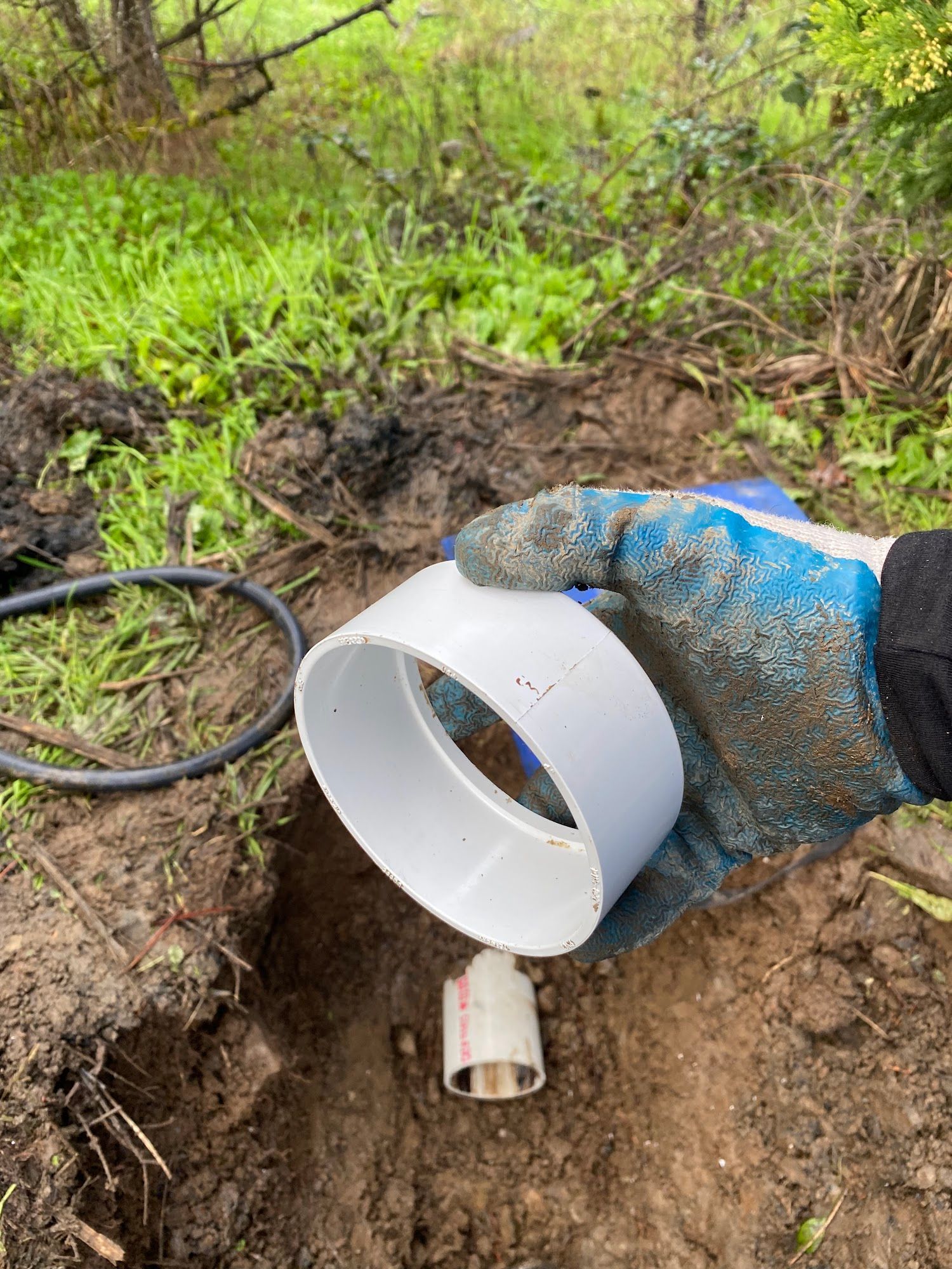 A gloved hand holds a white PVC pipe fitting above a hole in the dirt, where another pipe is visible.