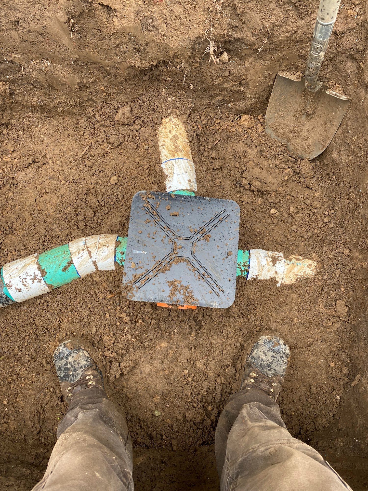 Person standing over a buried irrigation valve box with connected PVC pipes and a shovel nearby.