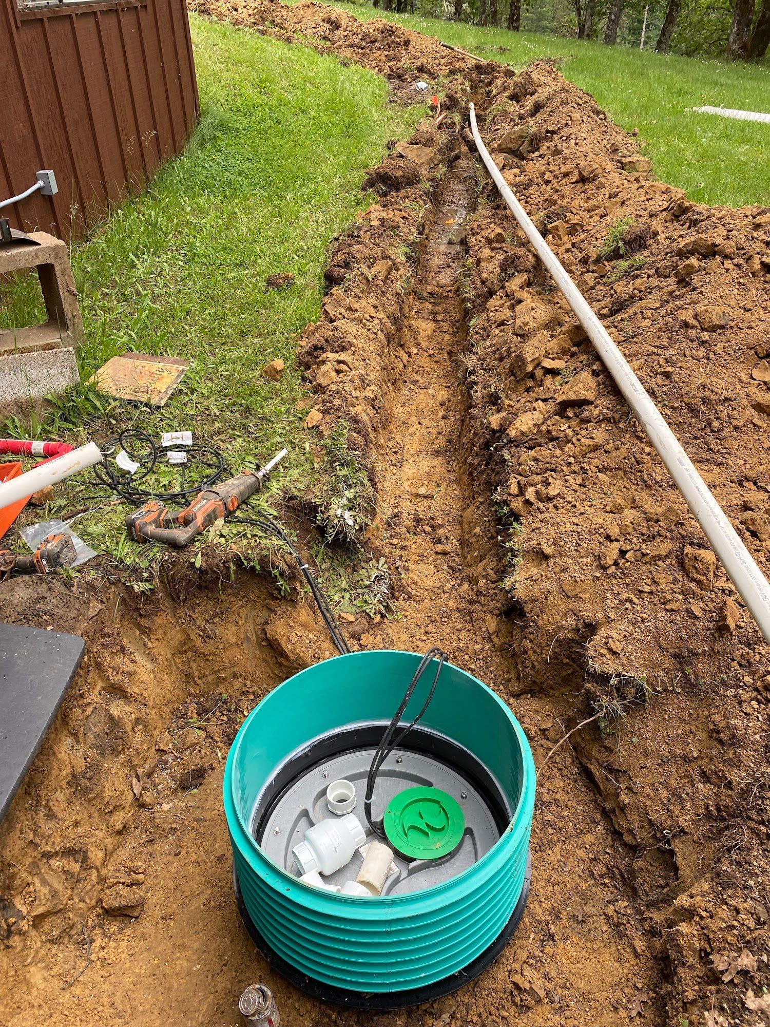 Green septic tank in trench, white pipe extending away, tools and disturbed earth.