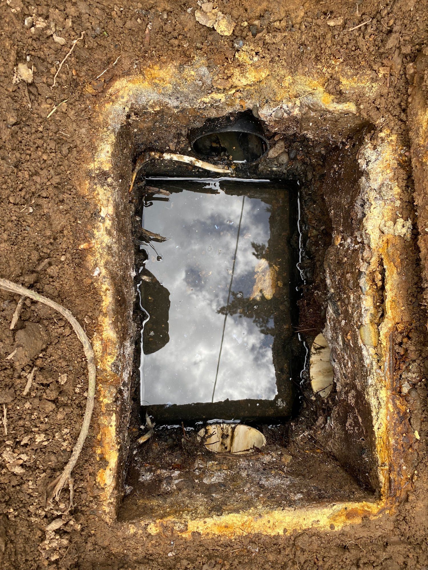 Excavated rectangular hole filled with water, reflecting cloudy sky. Black pipe visible.