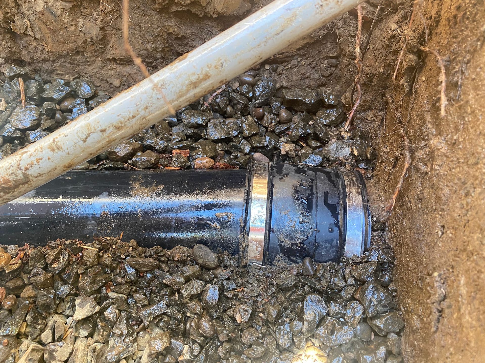 Black pipe connection in a gravel-filled trench with a gray pipe above.