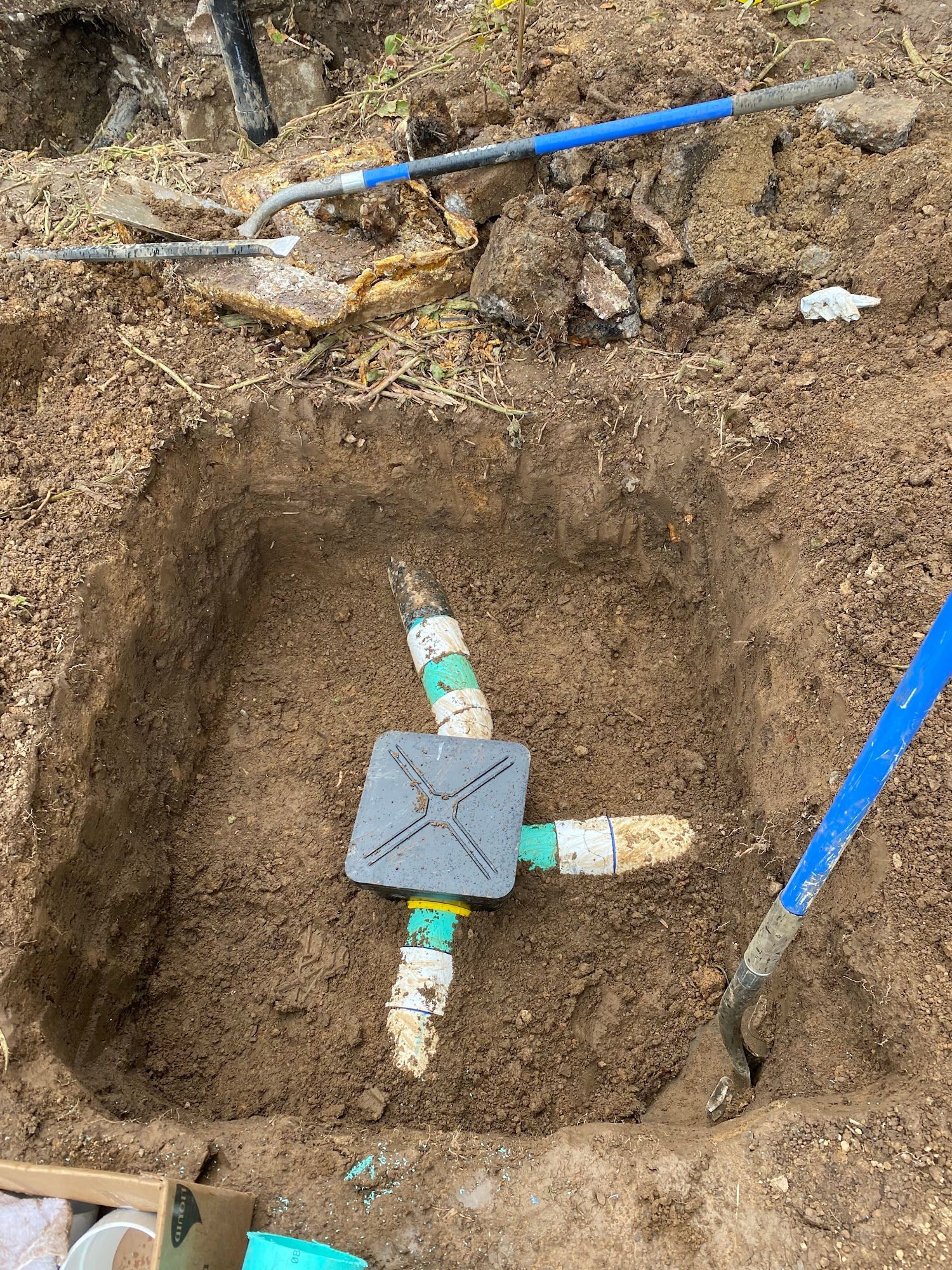 A square hole in the ground with a gray irrigation valve and white PVC pipes. Blue shovels and a rake lay nearby.