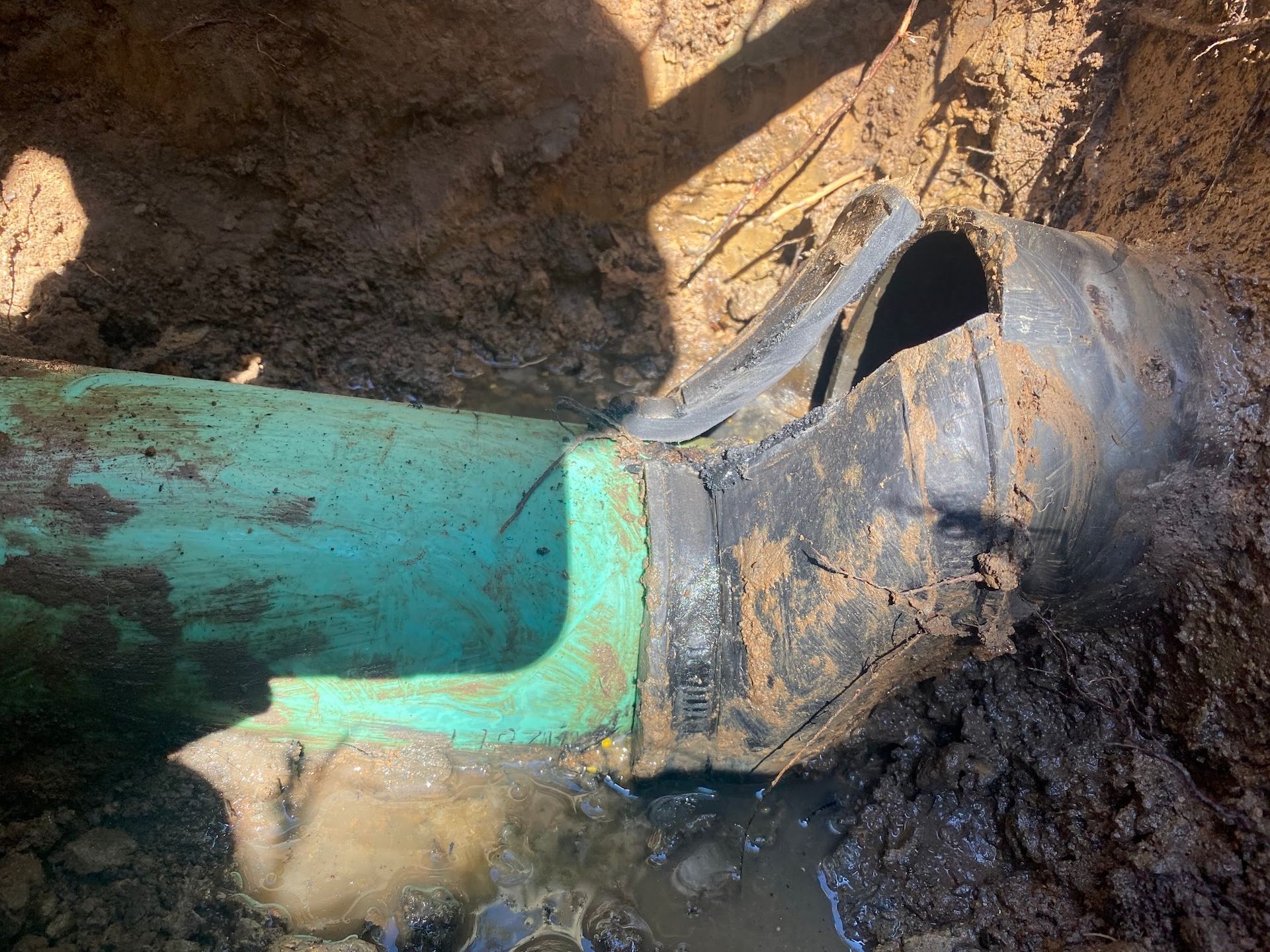 Green and black pipe connection exposed in muddy ground, possibly leaking water.