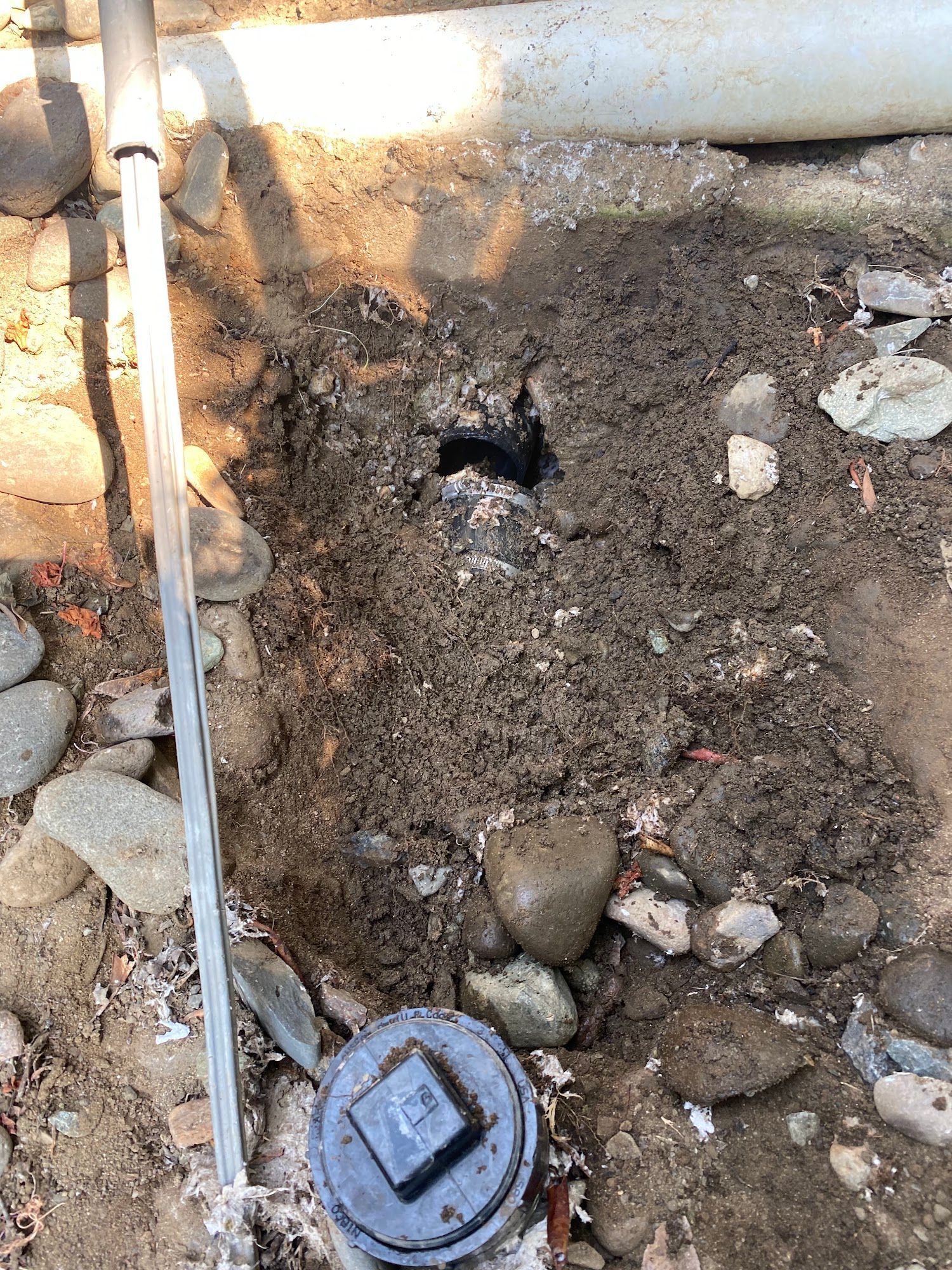 Hole in dirt with pipe visible, surrounded by rocks and a capped pipe access.