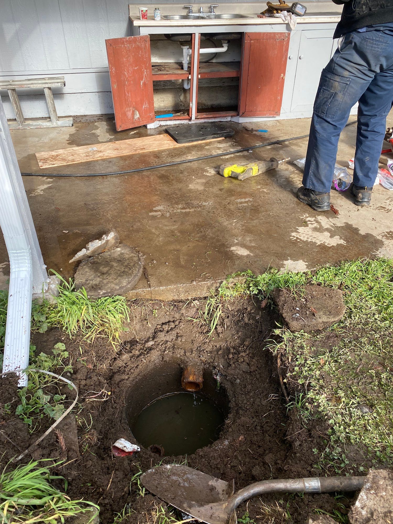 Open hole in ground with water, person in blue jeans, red cabinet, concrete, and grass.