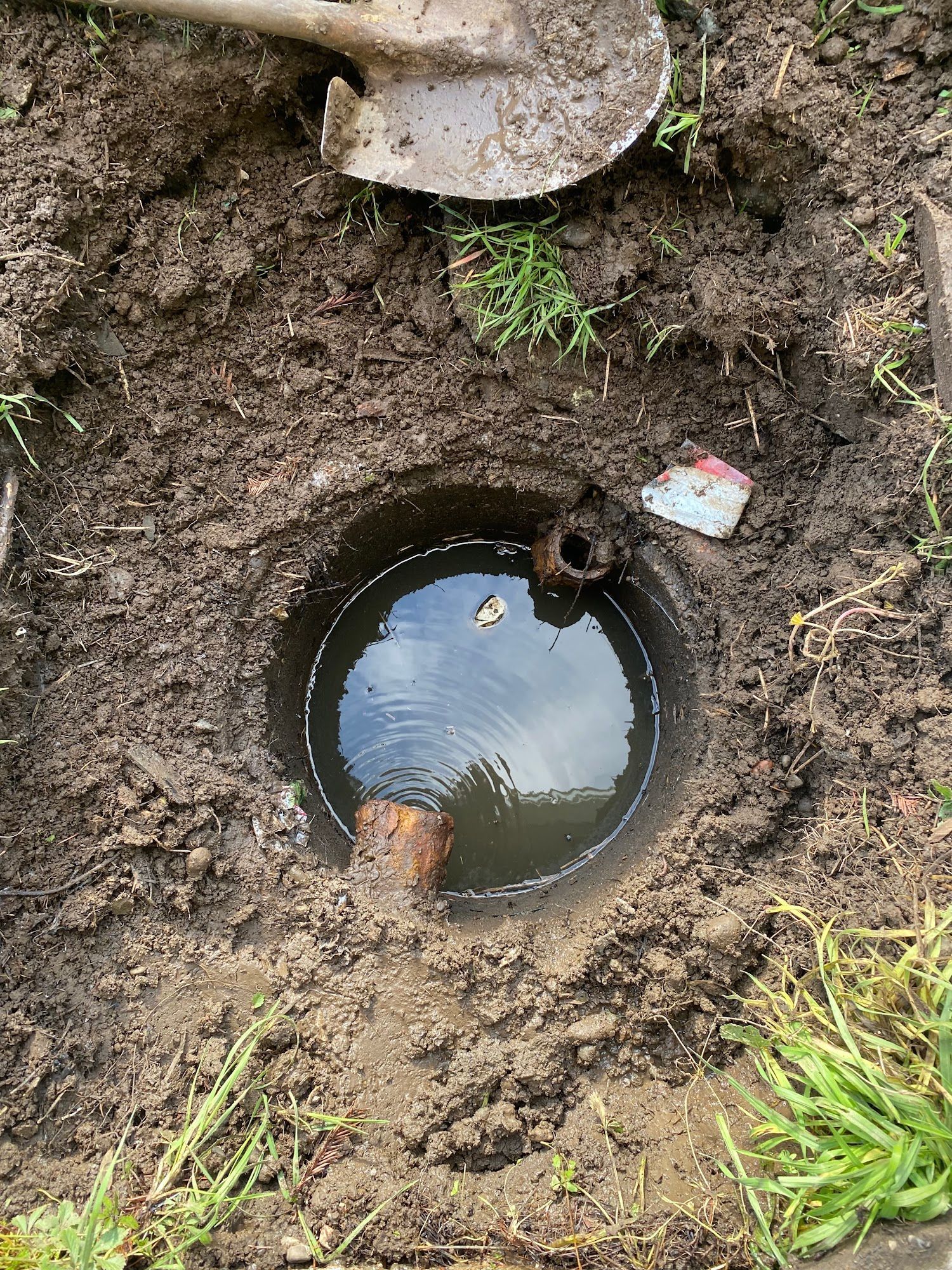 Hole dug in dirt, filled with water, revealing pipes. A shovel rests nearby.