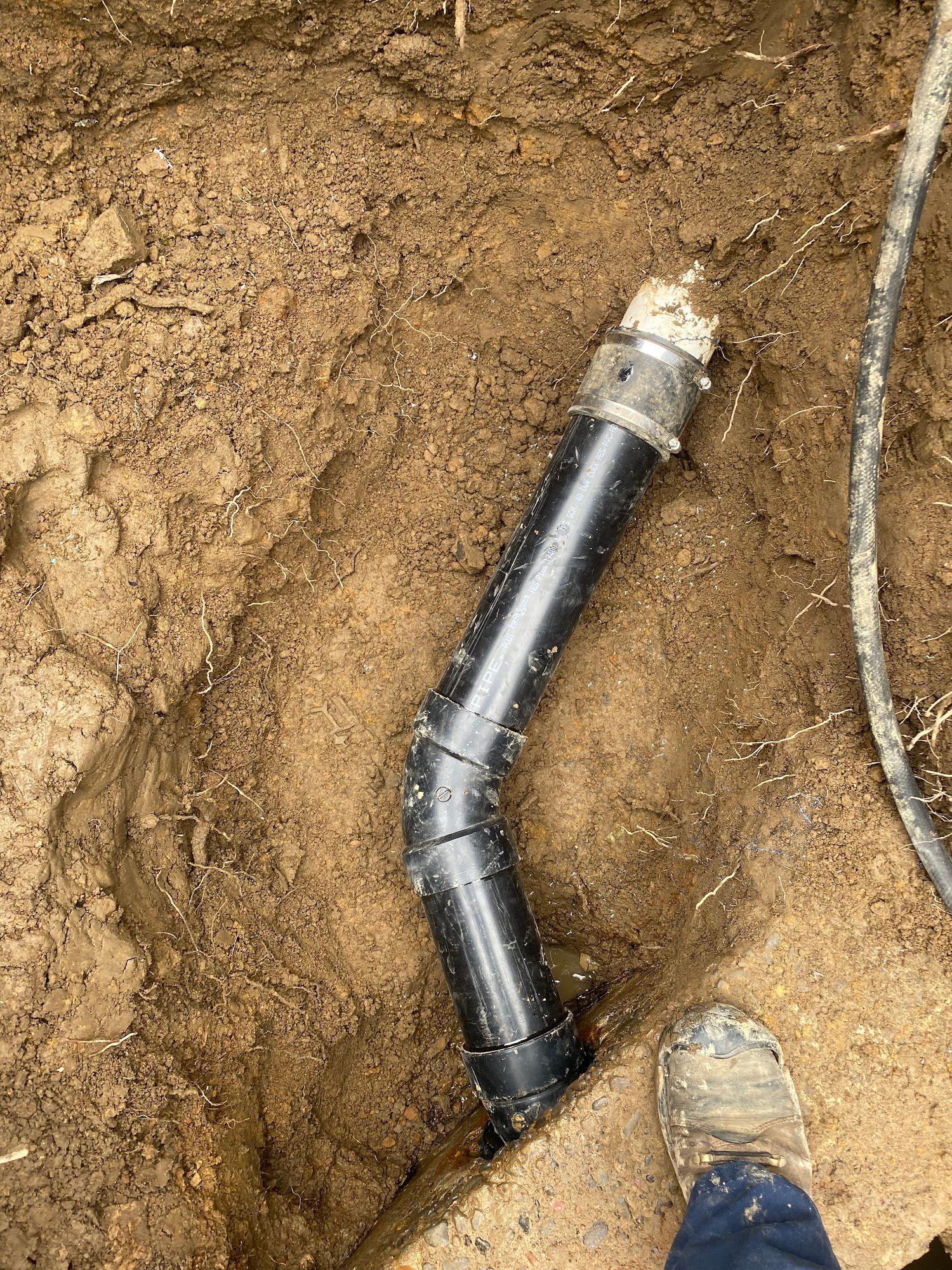 Black plumbing pipe in a dirt trench, connecting with a white fitting. A foot is visible.