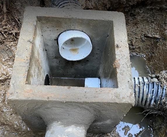Concrete drainage box with attached pipes, set in muddy soil.