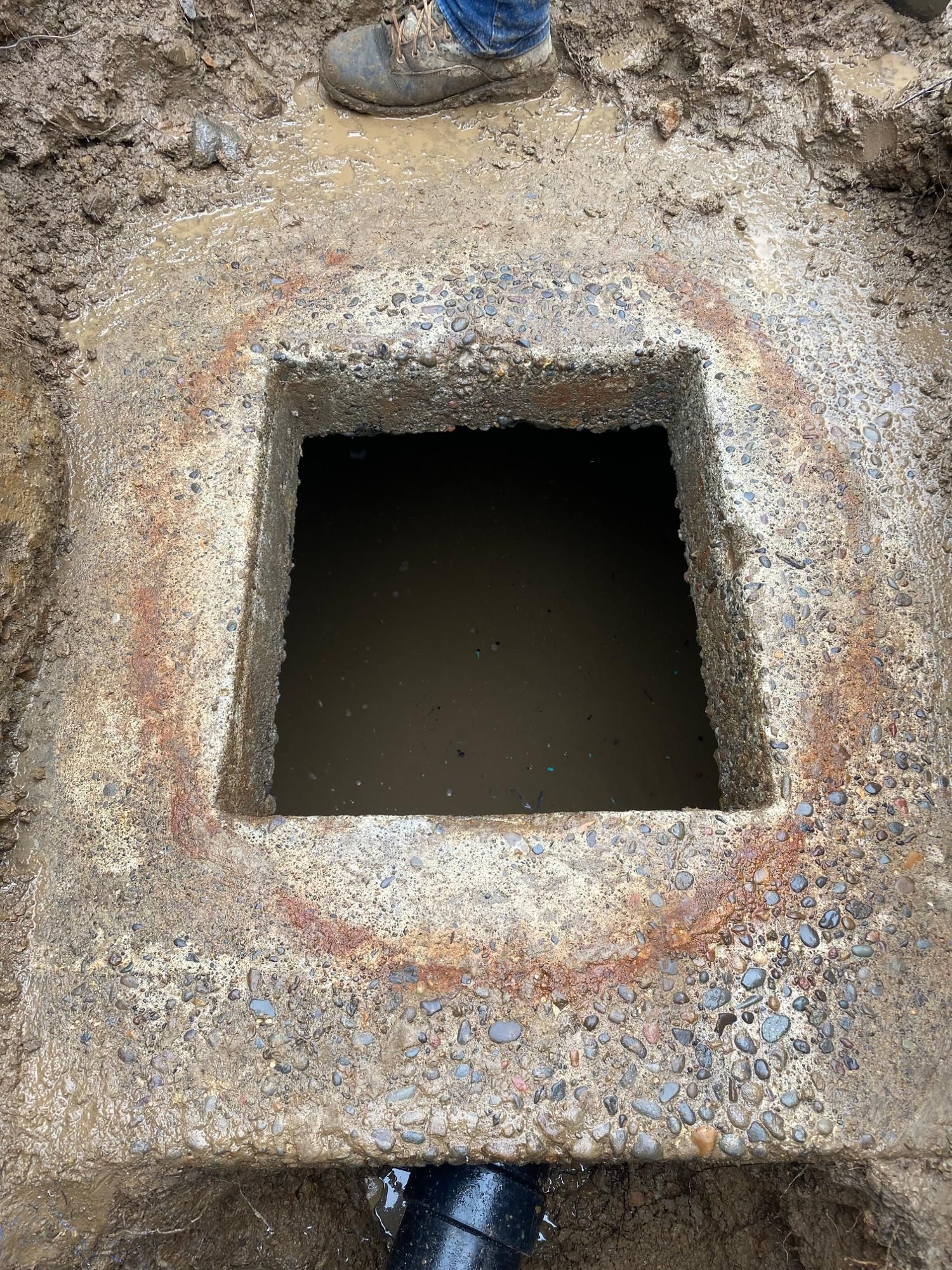 Open square hole in concrete, water visible inside. A person's boot stands at the top edge.