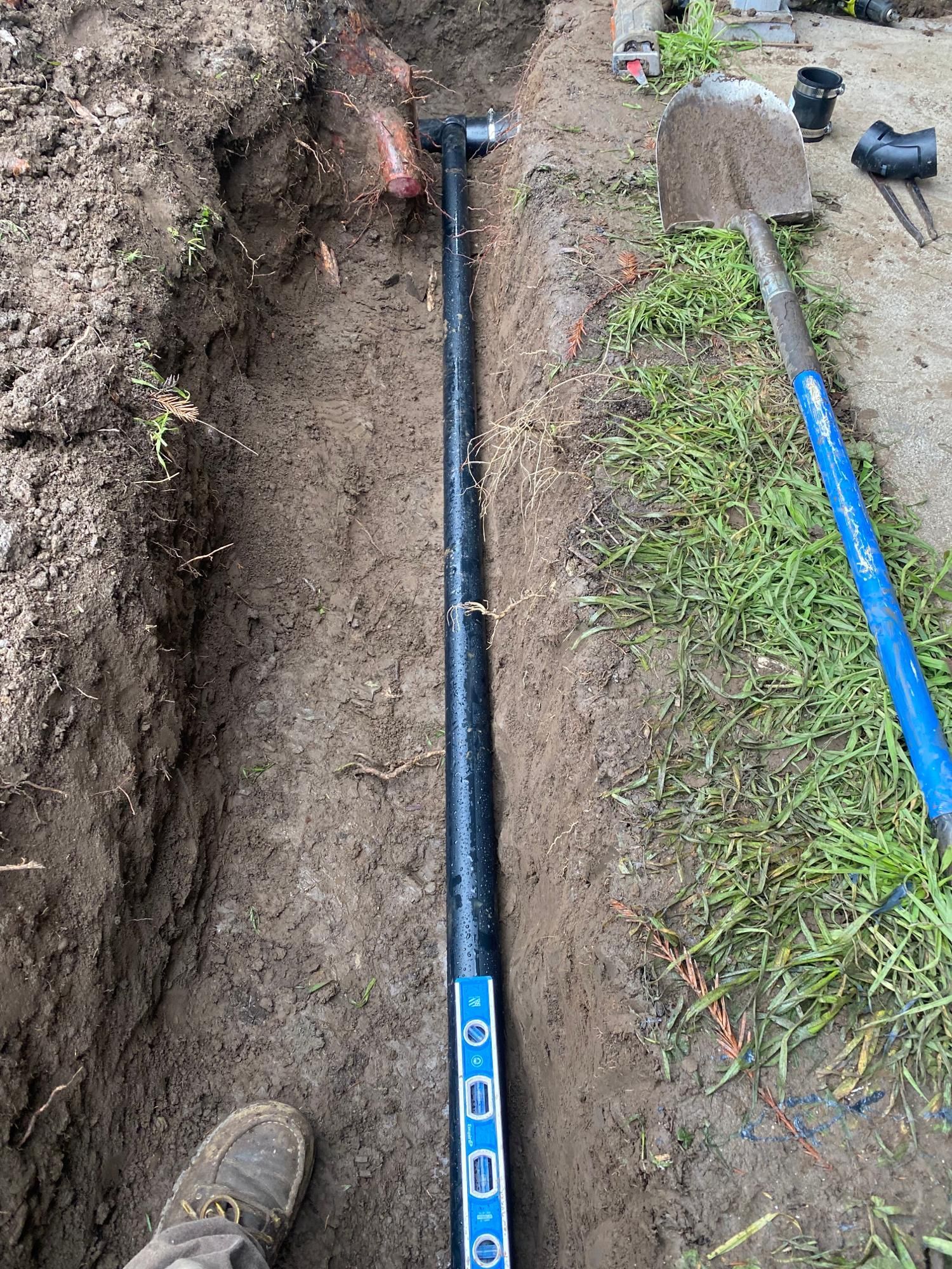 Trench with black pipe, level, shovel, and dirt, likely for a plumbing project outdoors.