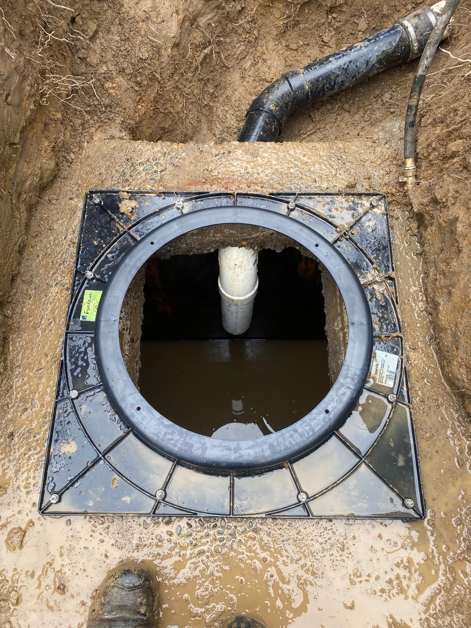 Square black cover with round opening in a dirt pit, showing a pipe and murky water.