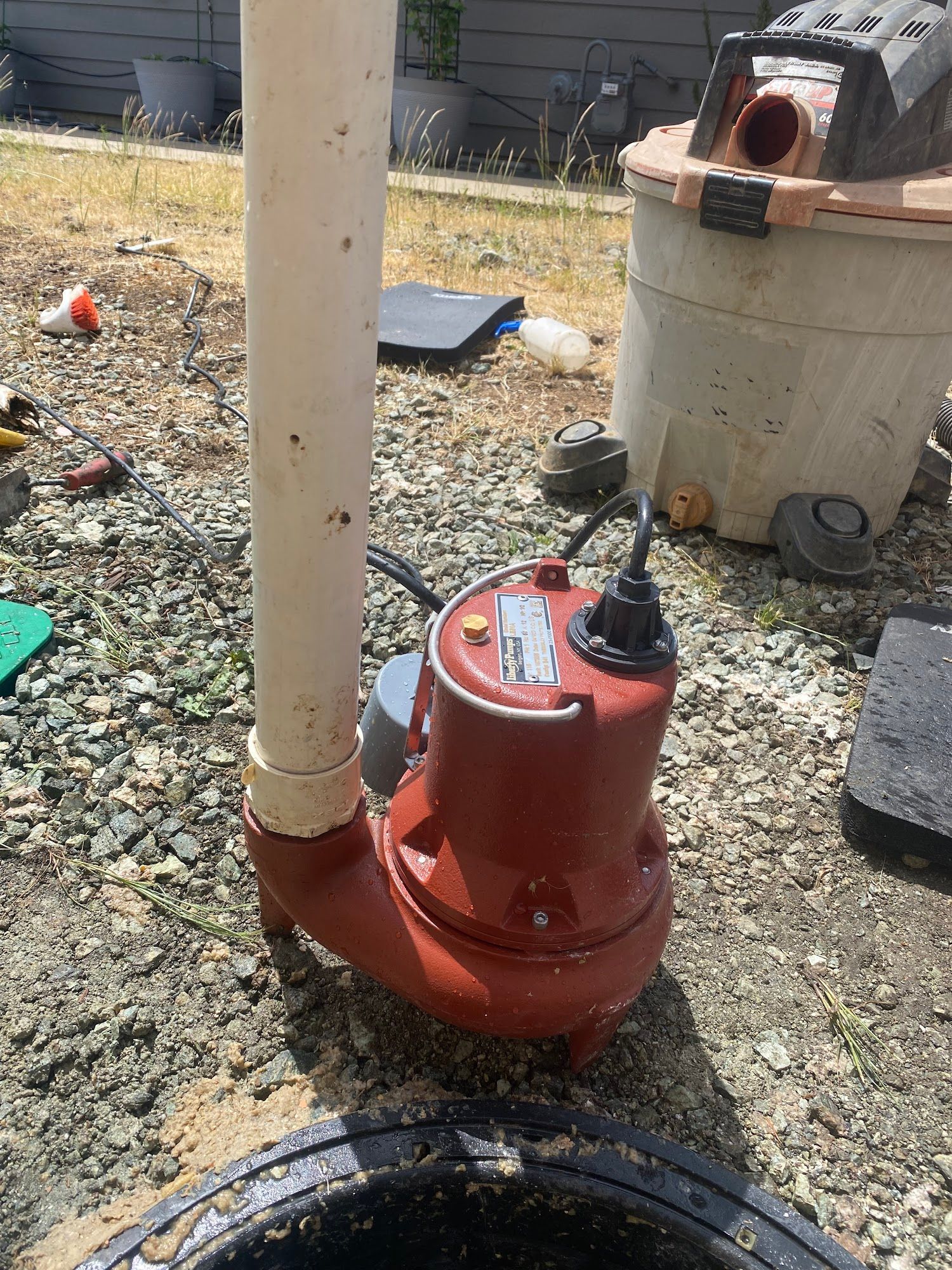 Red submersible pump with white pipe in a dark round opening, outdoors.