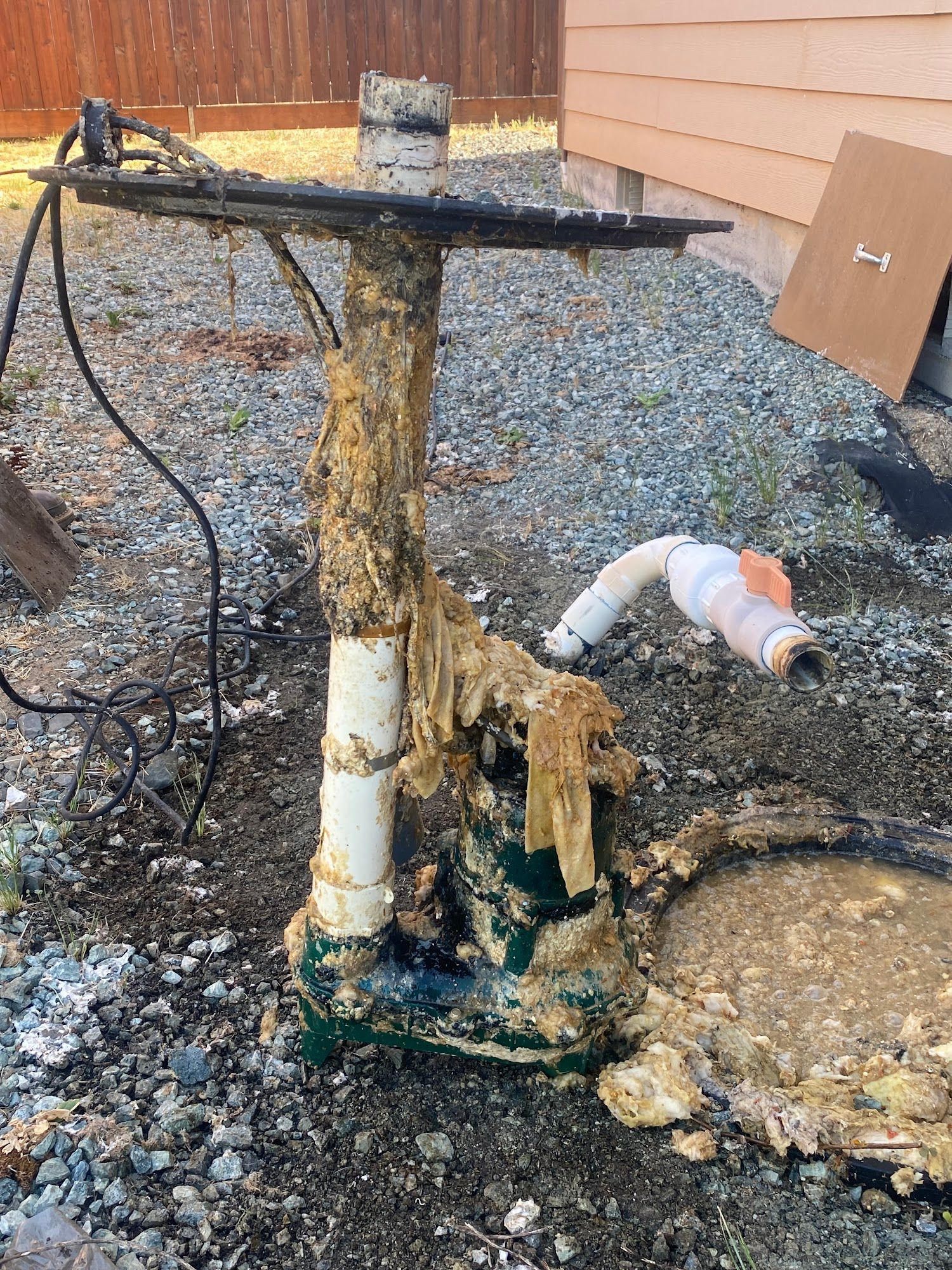 A dirty sewage pump in a gravel yard, covered in sludge, with white pipe and electrical wiring.