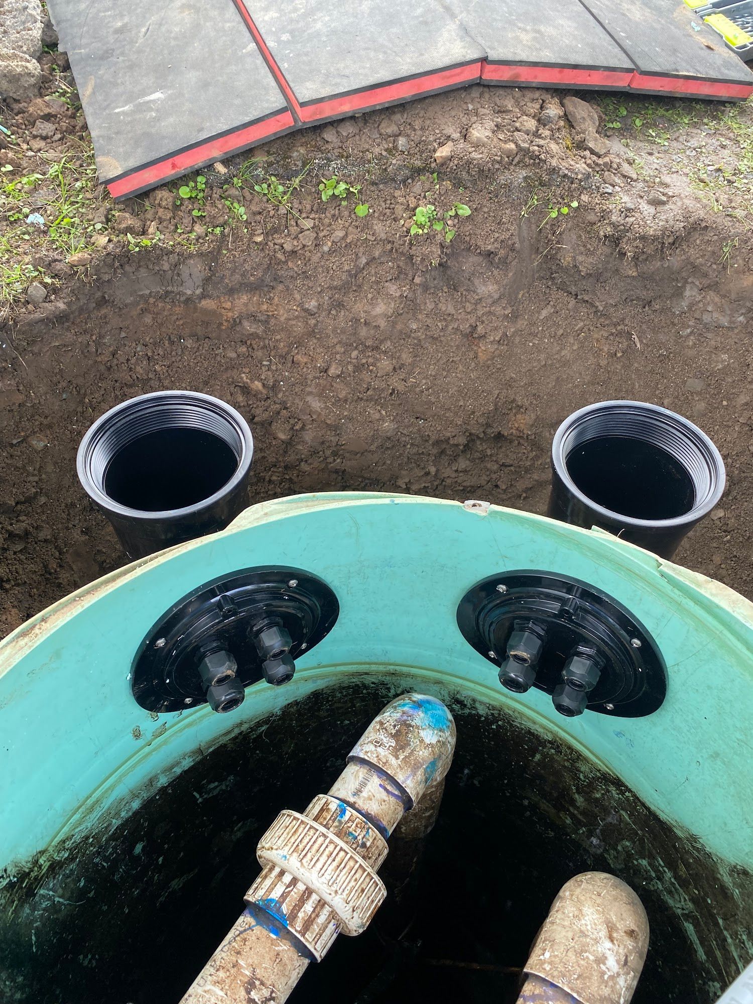Green septic tank with two black openings and pipes inside, surrounded by dirt and two open black cylinders.