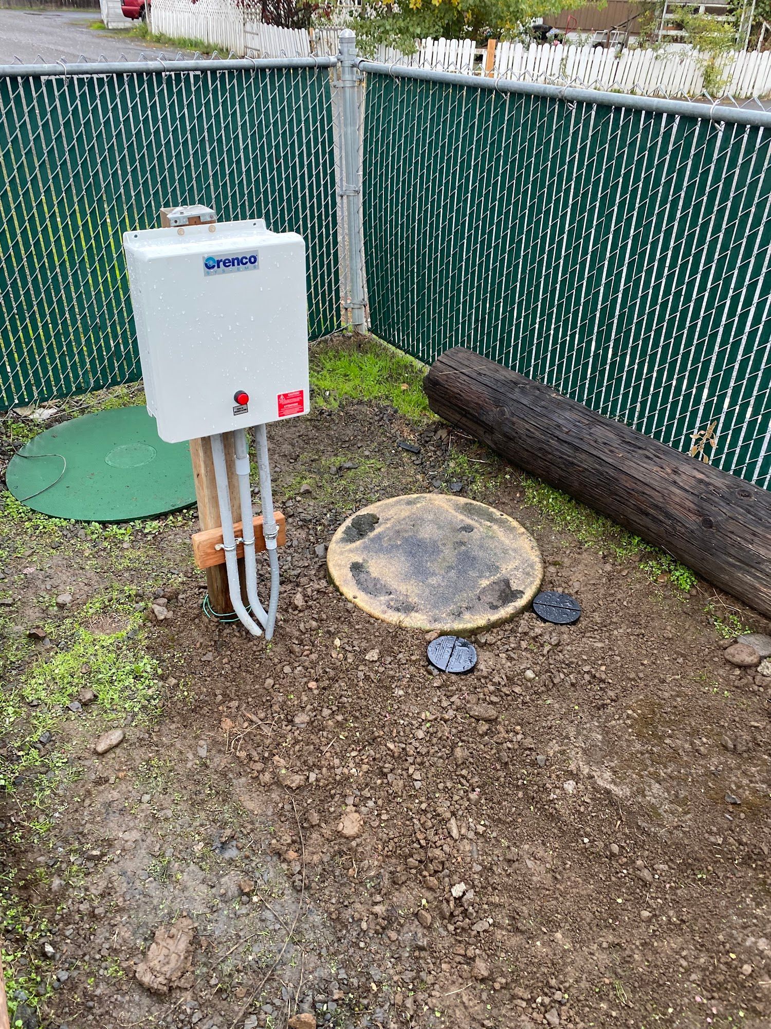 A tankless water heater, two access covers, and a fence in an outdoor setting.