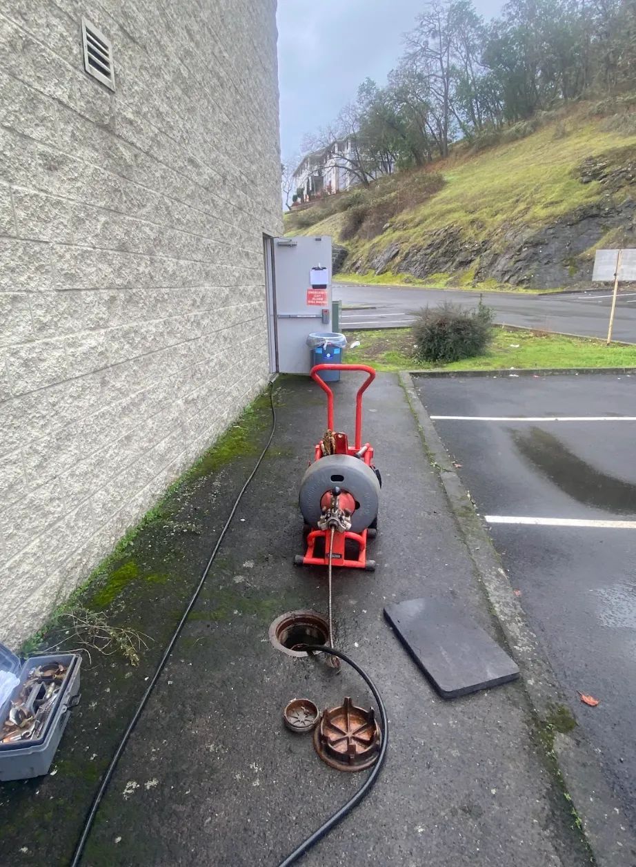 Plumbing equipment next to an open sewer access point on a paved surface near a building.