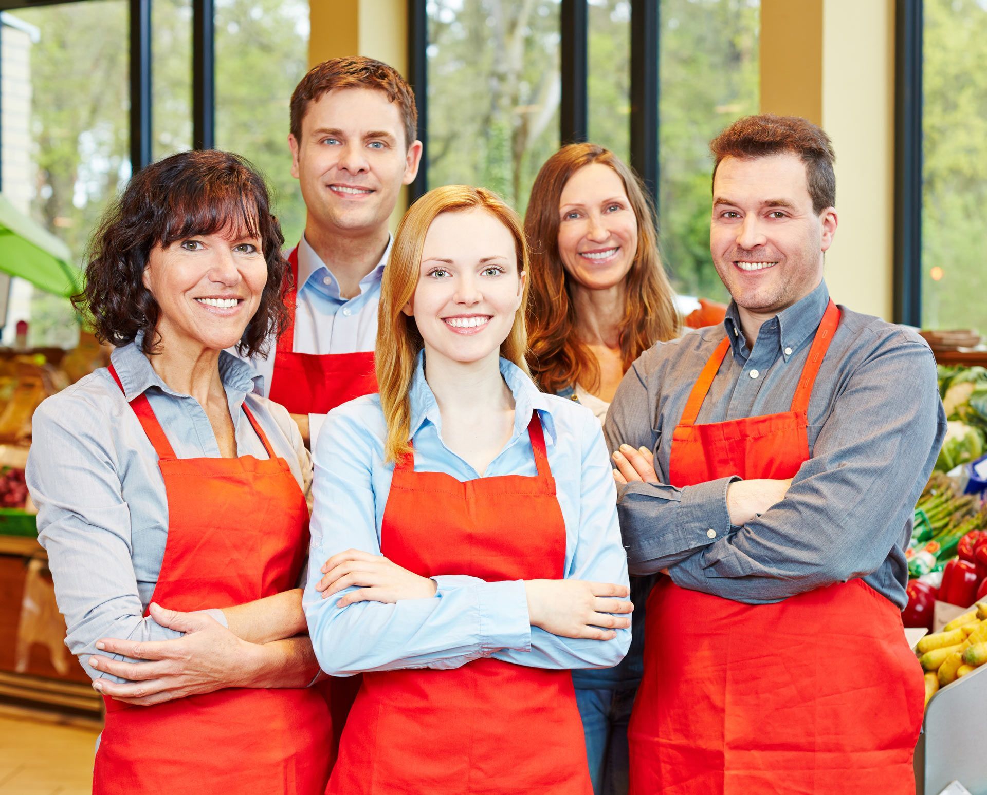 Five grocery store employees wearing red aprons, smiling, standing inside a store.