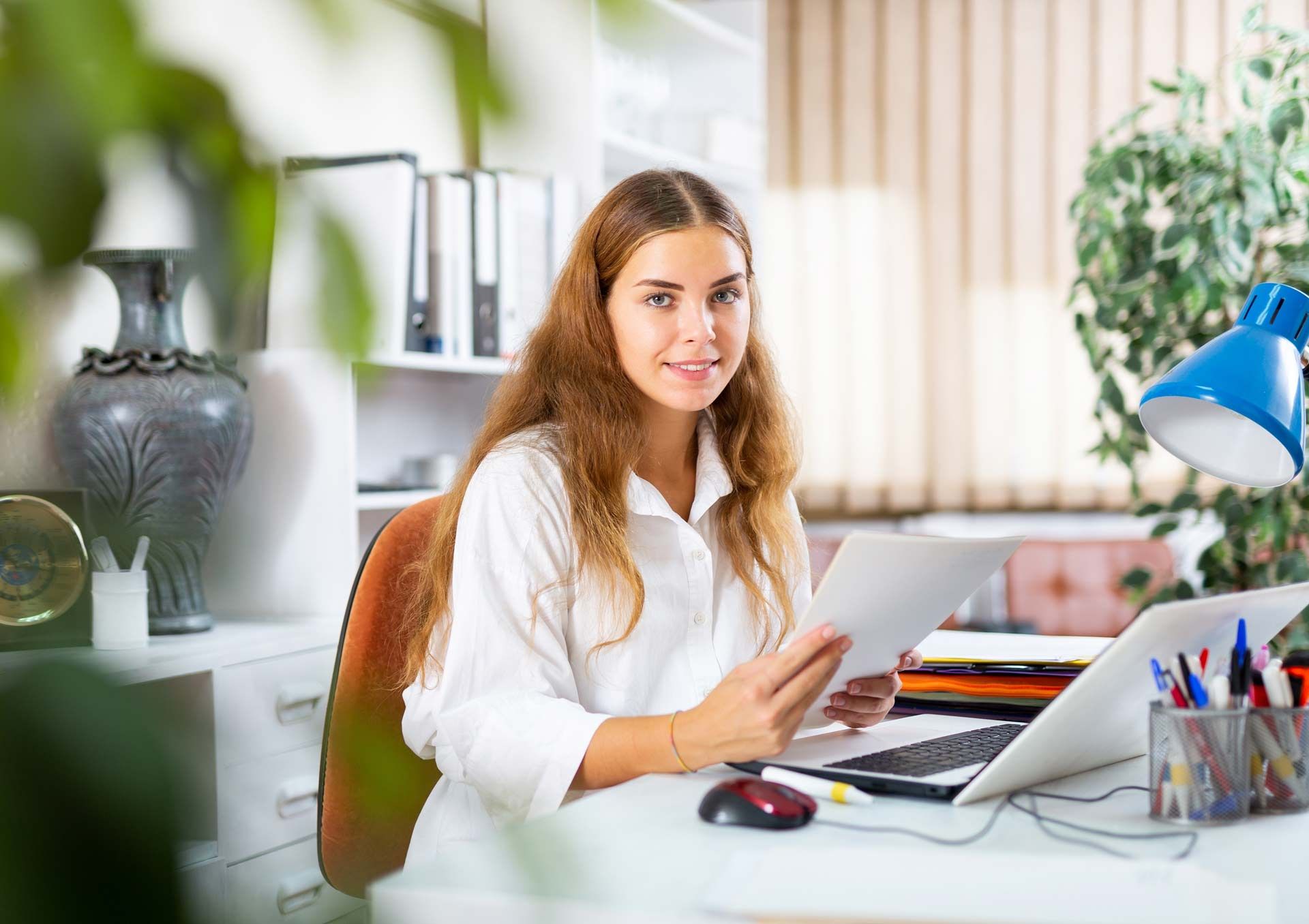 Woman in white shirt, holding papers, smiles while working at a desk with a laptop.