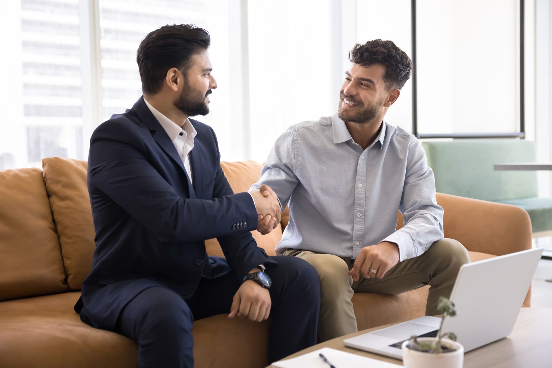 Two men shaking hands on a brown sofa, smiling.