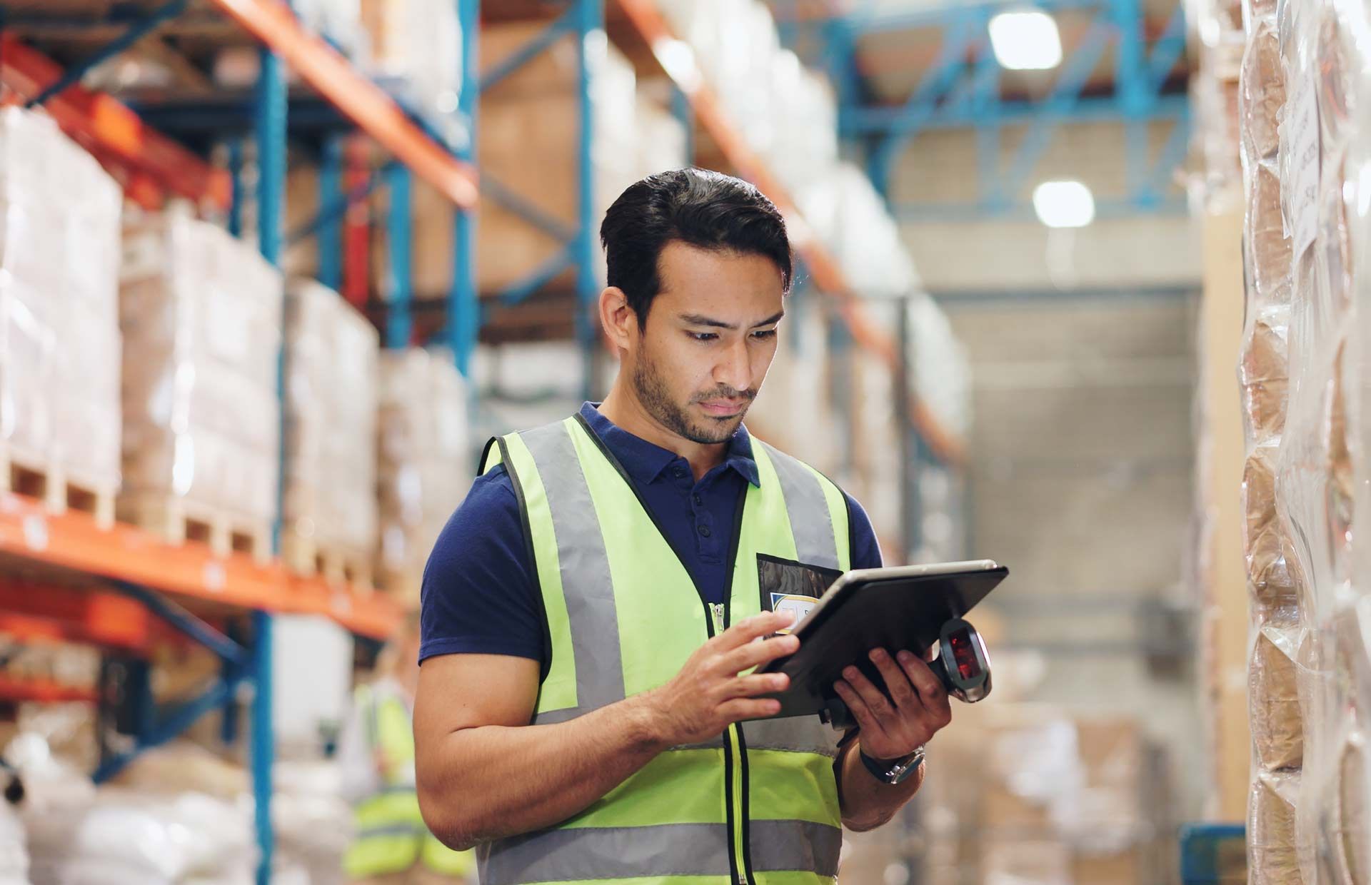 Man in a warehouse, wearing a safety vest, looking at a tablet.