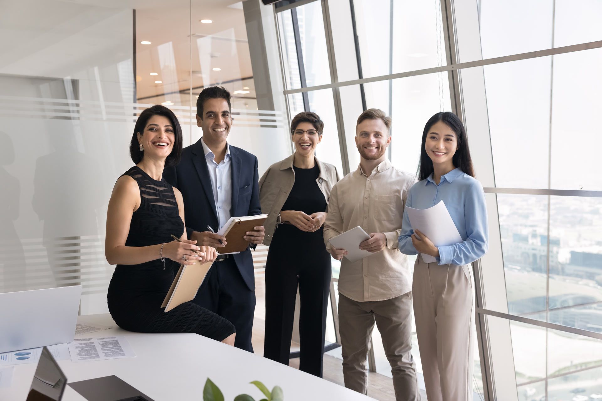 Group of five professionals smiling in a modern office with large windows; holding documents.