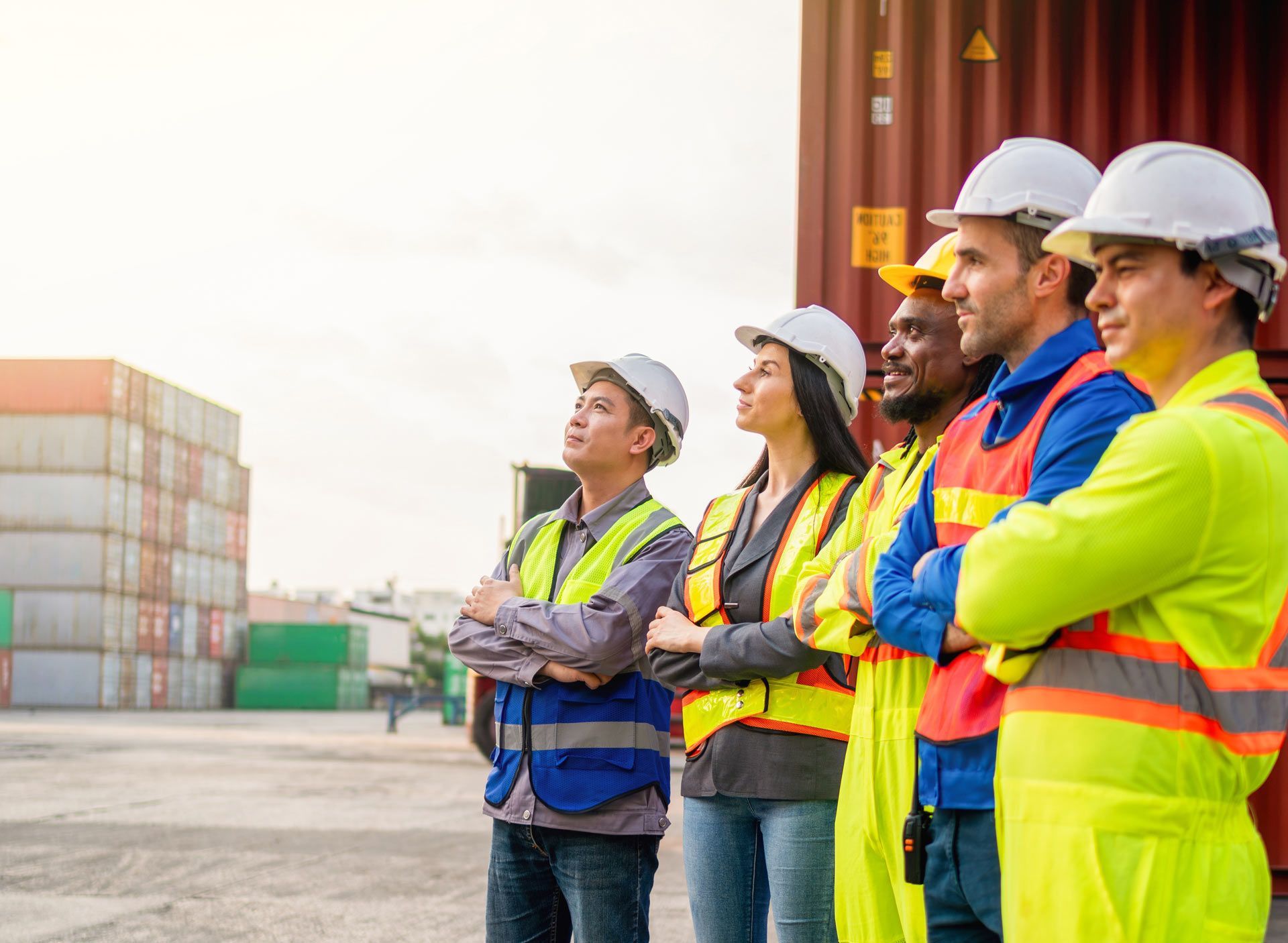 Workers in hard hats and safety vests stand near shipping containers, looking upward.