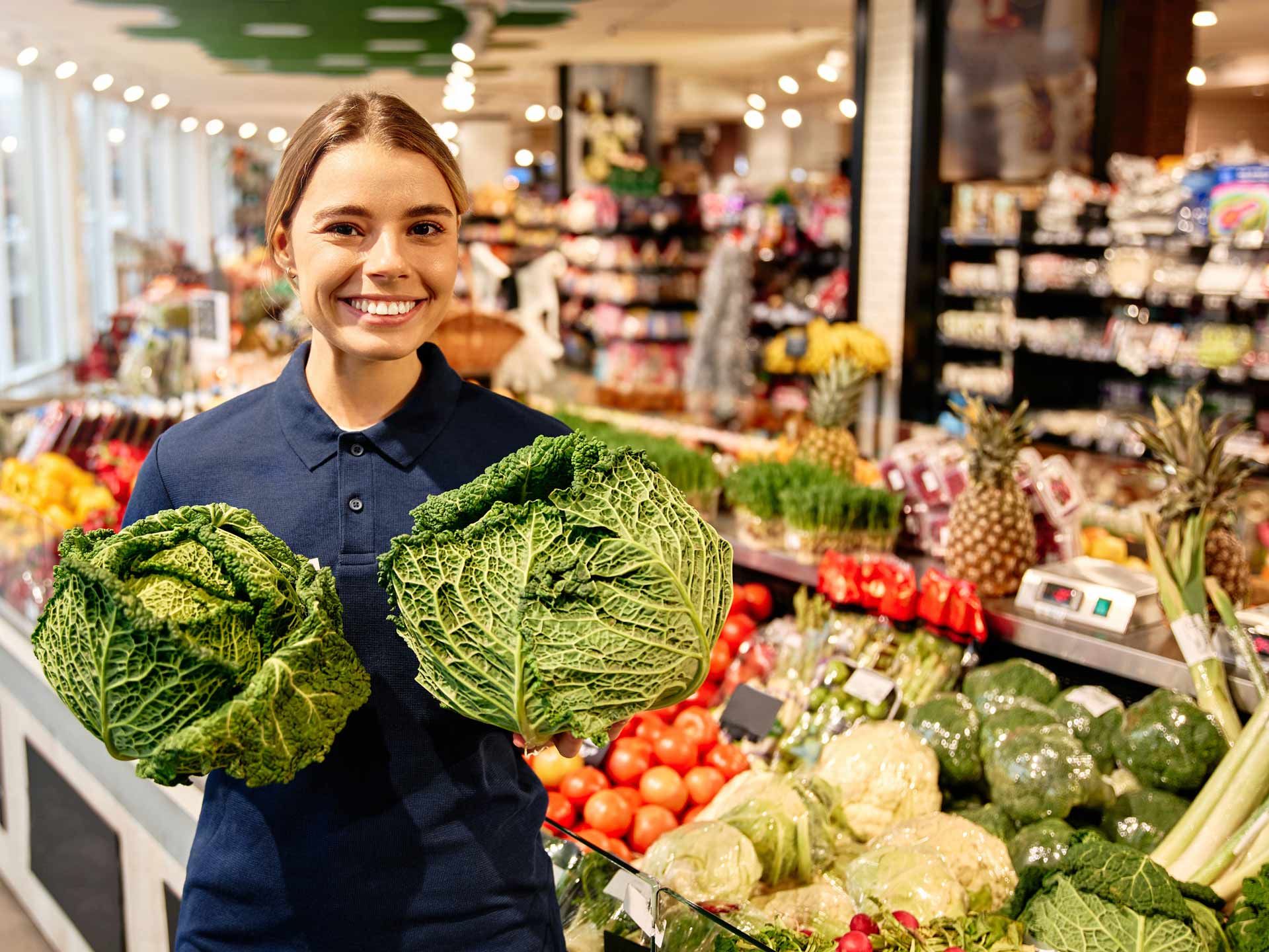 Woman in grocery store smiles, holding two green cabbages.