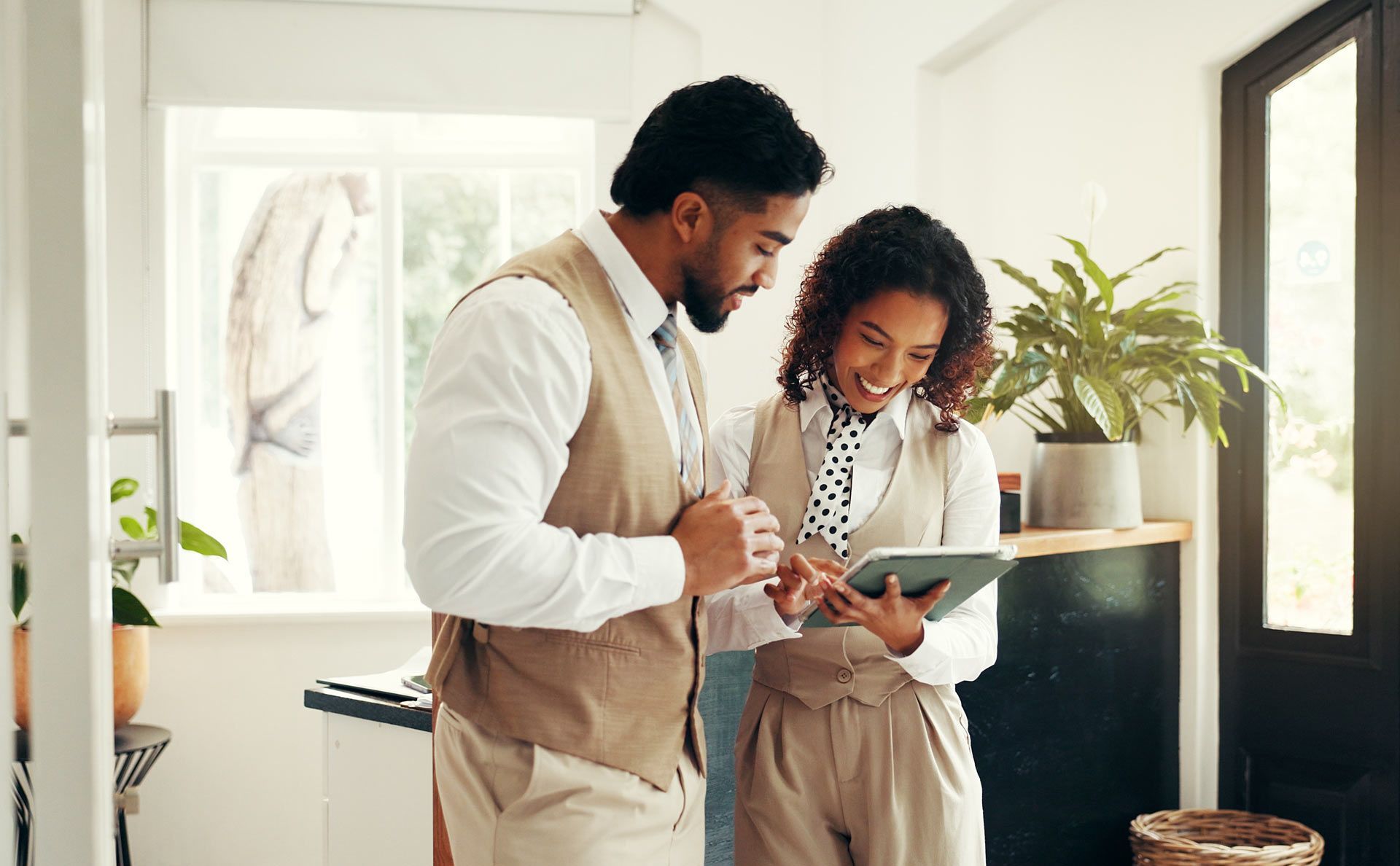 Two people in business attire look at a tablet near a window, smiling.