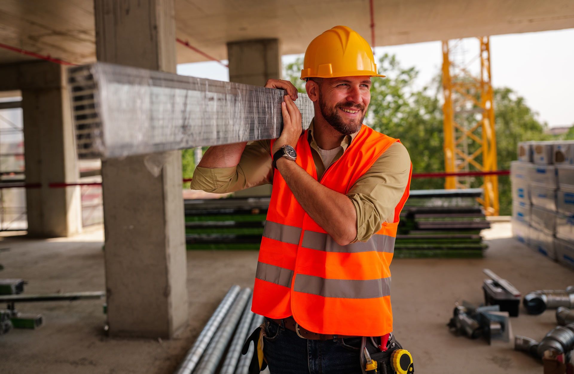 Construction worker carrying metal beam, wearing hard hat and safety vest, smiling on a construction site.
