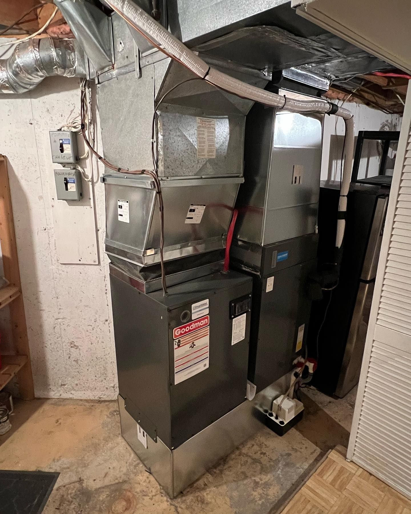 An air conditioner is sitting in a basement next to a refrigerator.