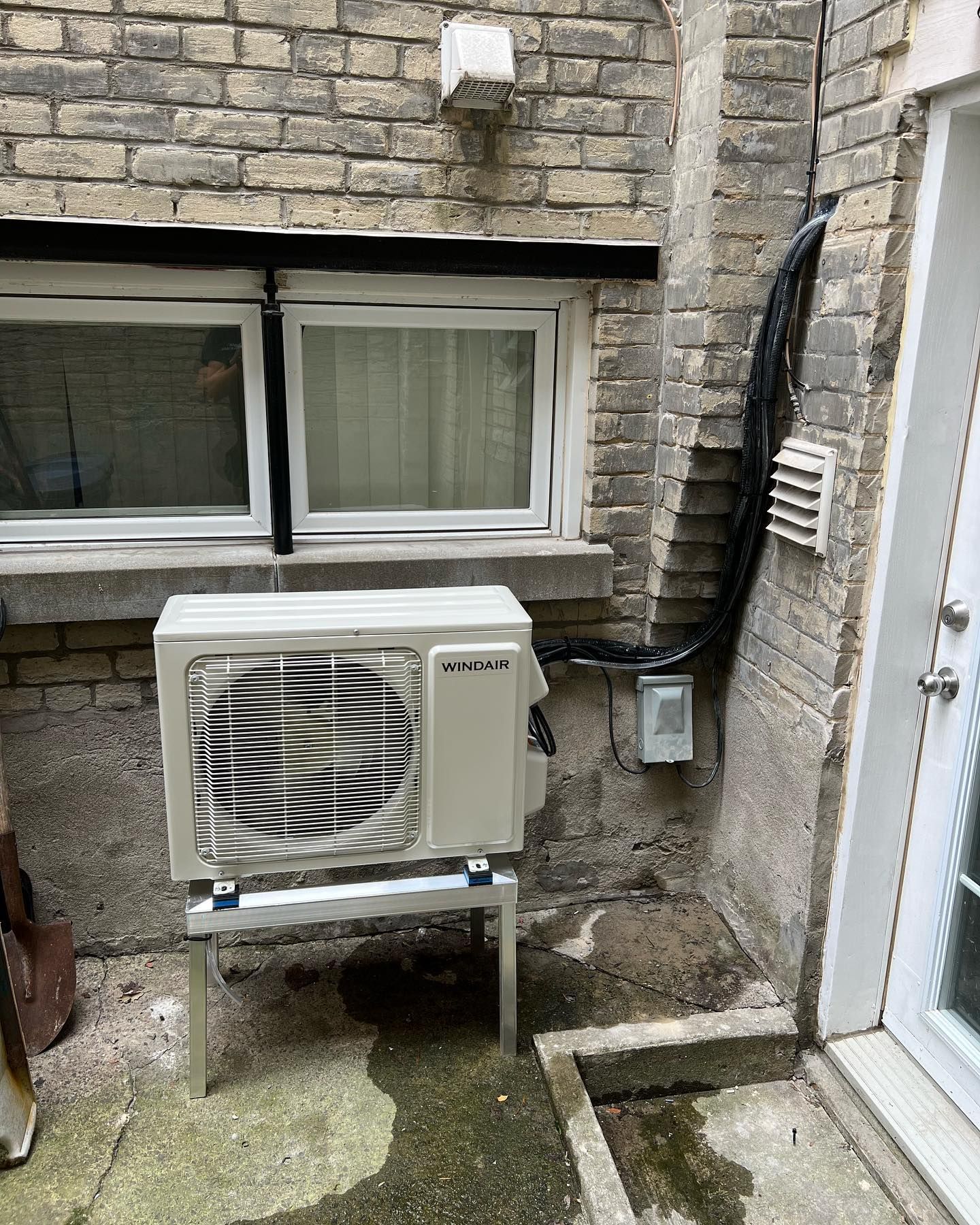 A white air conditioner is sitting outside of a brick building next to a window.