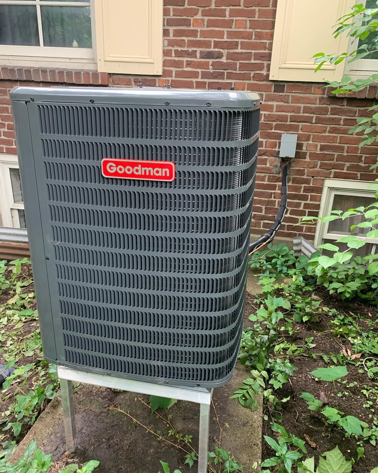 A goodman air conditioner is sitting outside of a brick building.
