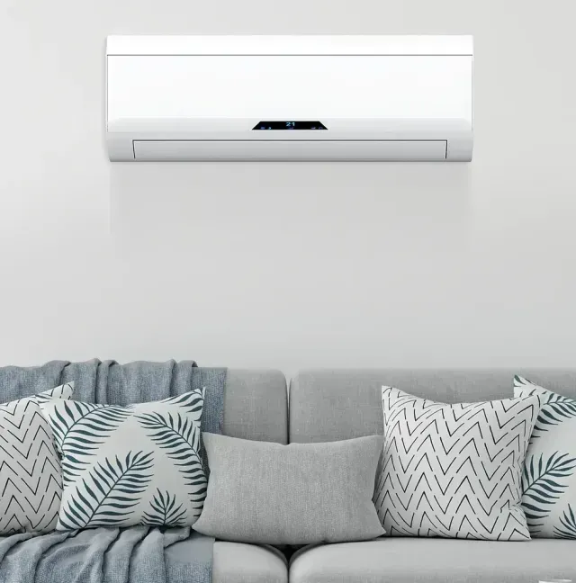 Un climatiseur blanc au-dessus d'un canapé gris avec des coussins et un plaid bleu, sur un mur blanc.