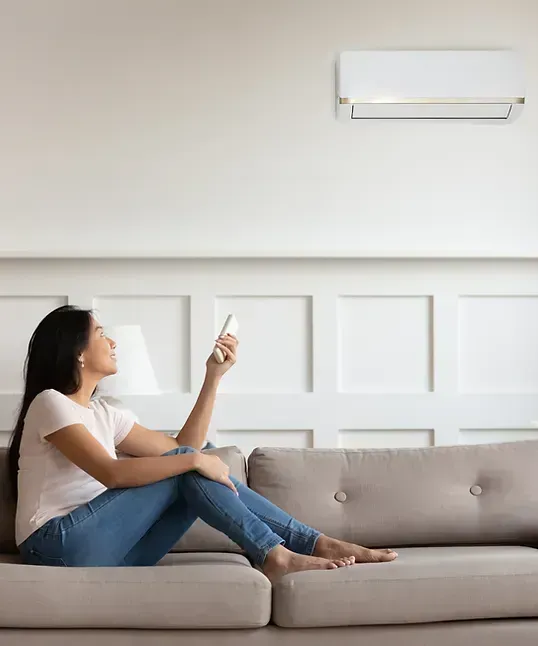 Une femme assise sur un canapé règle un climatiseur mural à l'aide d'une télécommande.